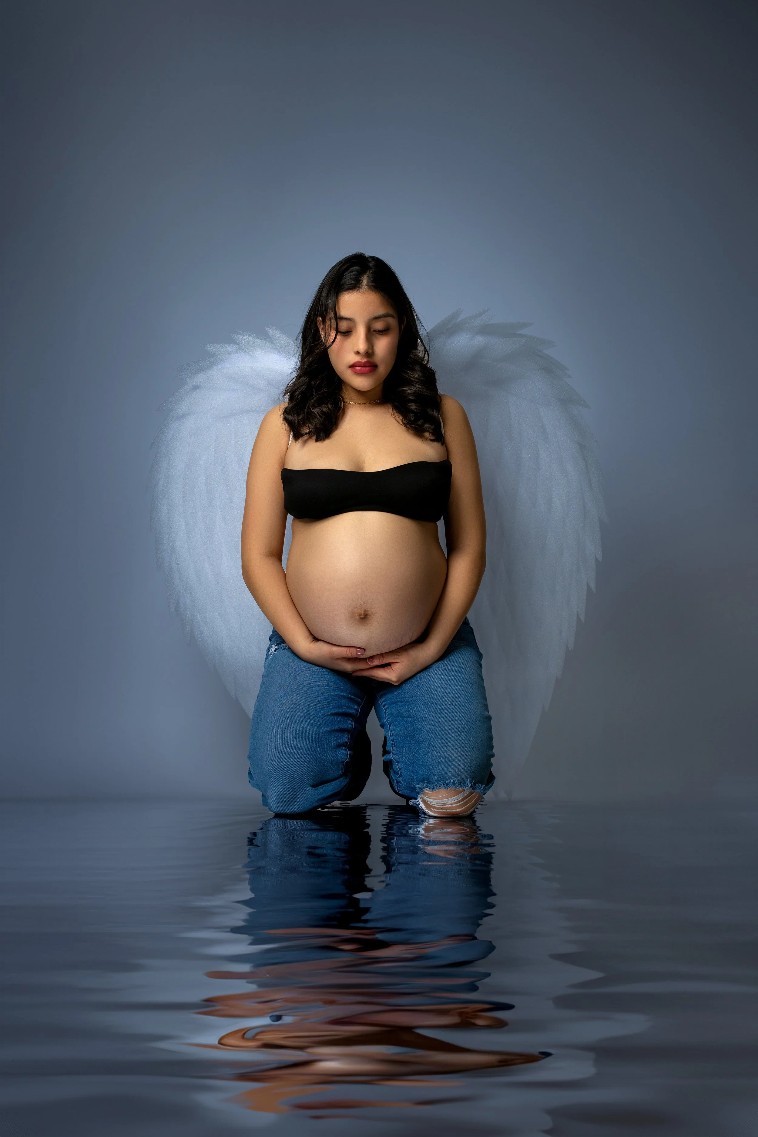 A pregnant woman with dark hair and closed eyes, wearing a black tube top, blue jeans, and angel wings, kneeling on water with a reflective surface.