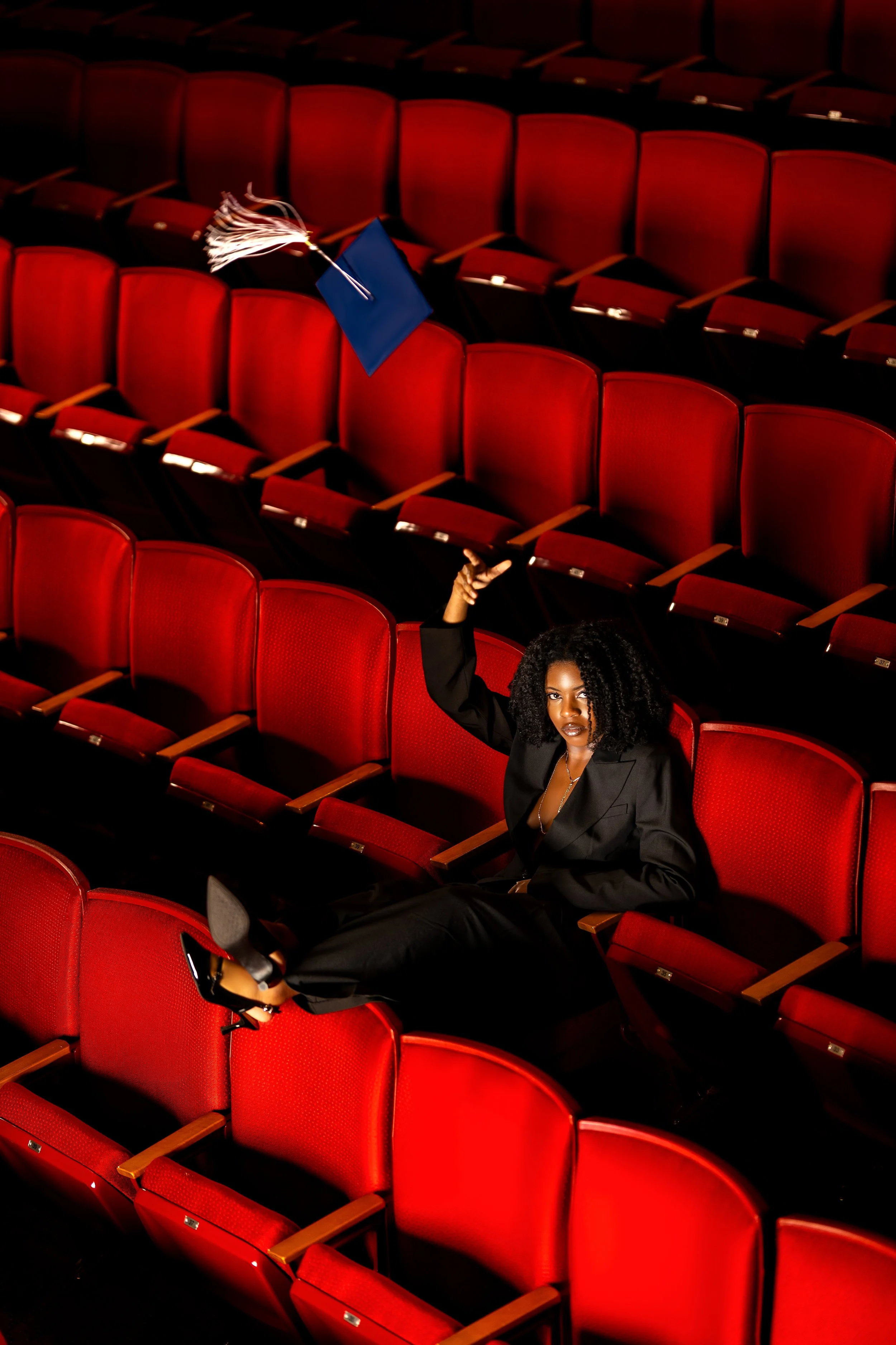 A woman sitting in an empty theater with rows of red seats. She is dressed in a black outfit, resting her arm on a seat, and holding a diploma in her other hand while looking at the camera. A blue graduation cap with a tassel is placed on an empty seat.