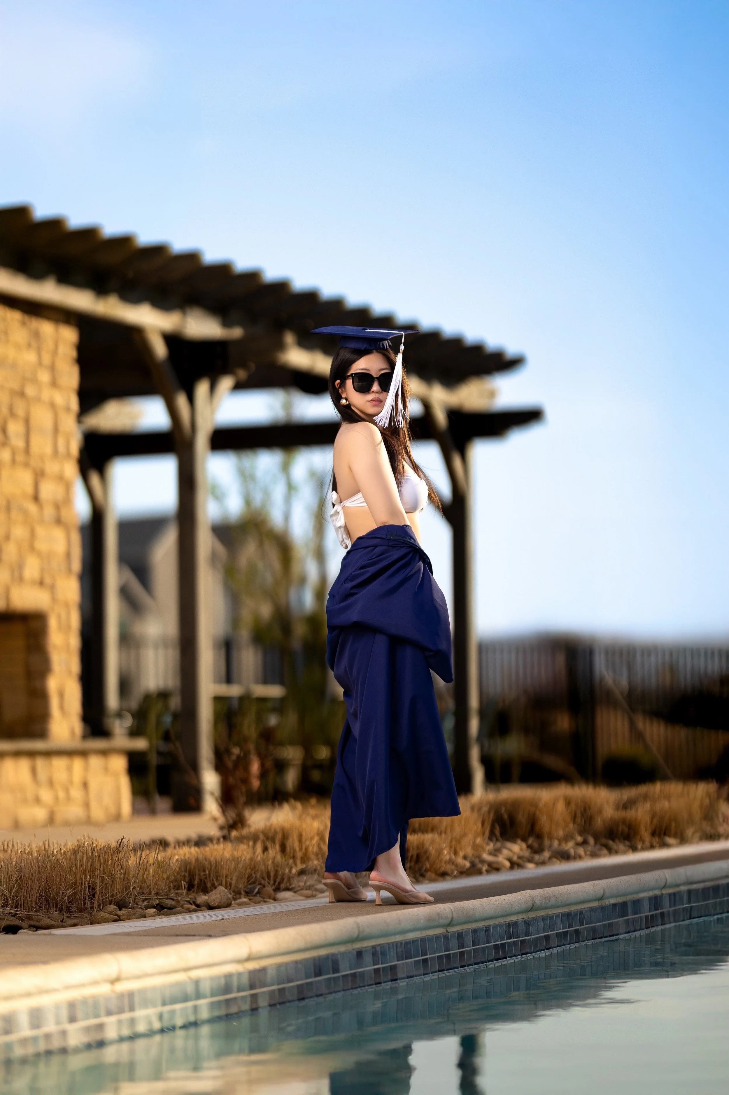 A woman wearing sunglasses, a white top, a navy blue skirt, and nude heels, standing near a swimming pool with a graduation cap on her head. She is looking over her shoulder, with a backdrop of a wooden pergola and clear blue sky.