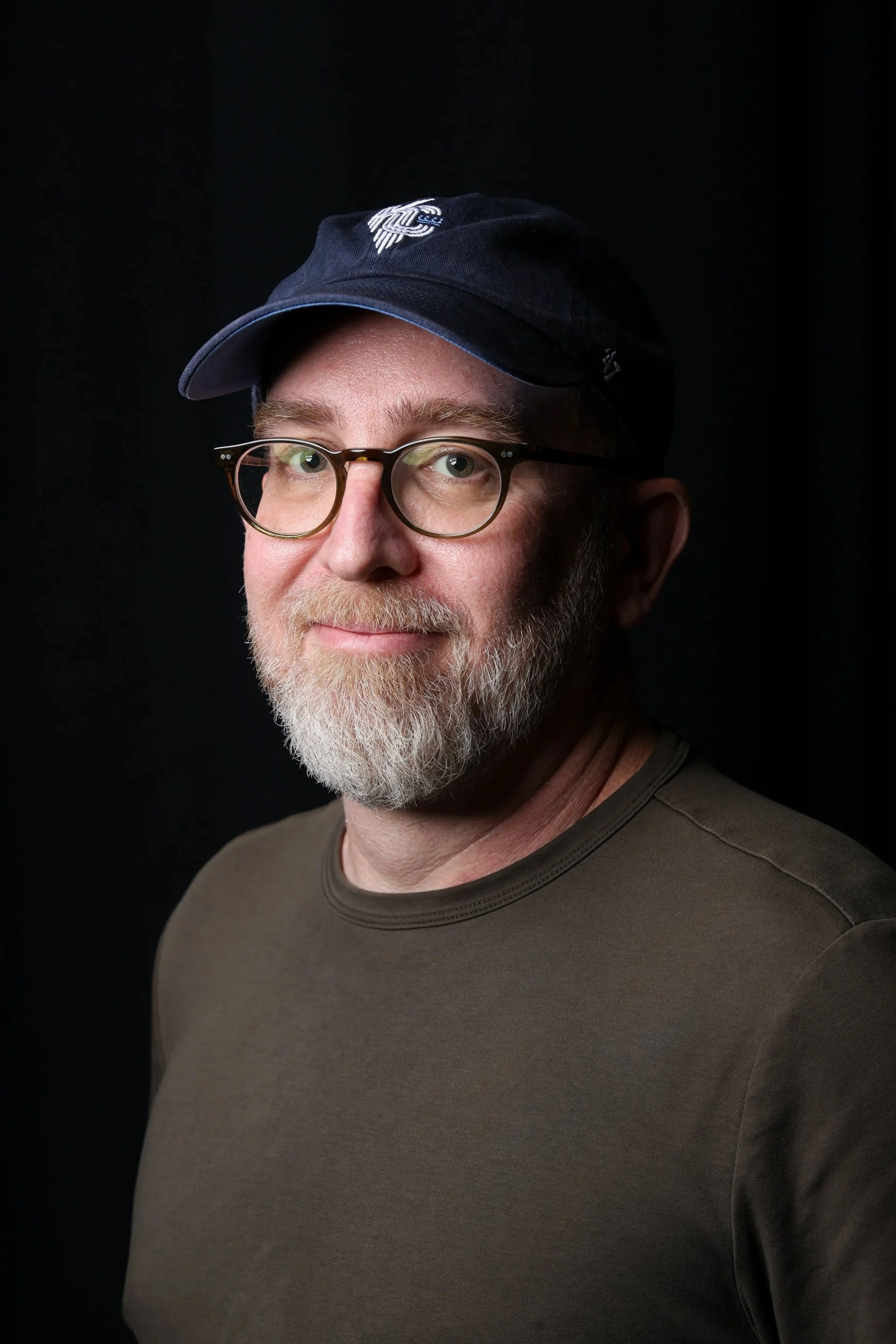 Close-up portrait of a mature man with a white beard and glasses, wearing a dark baseball cap with a logo, against a black background.