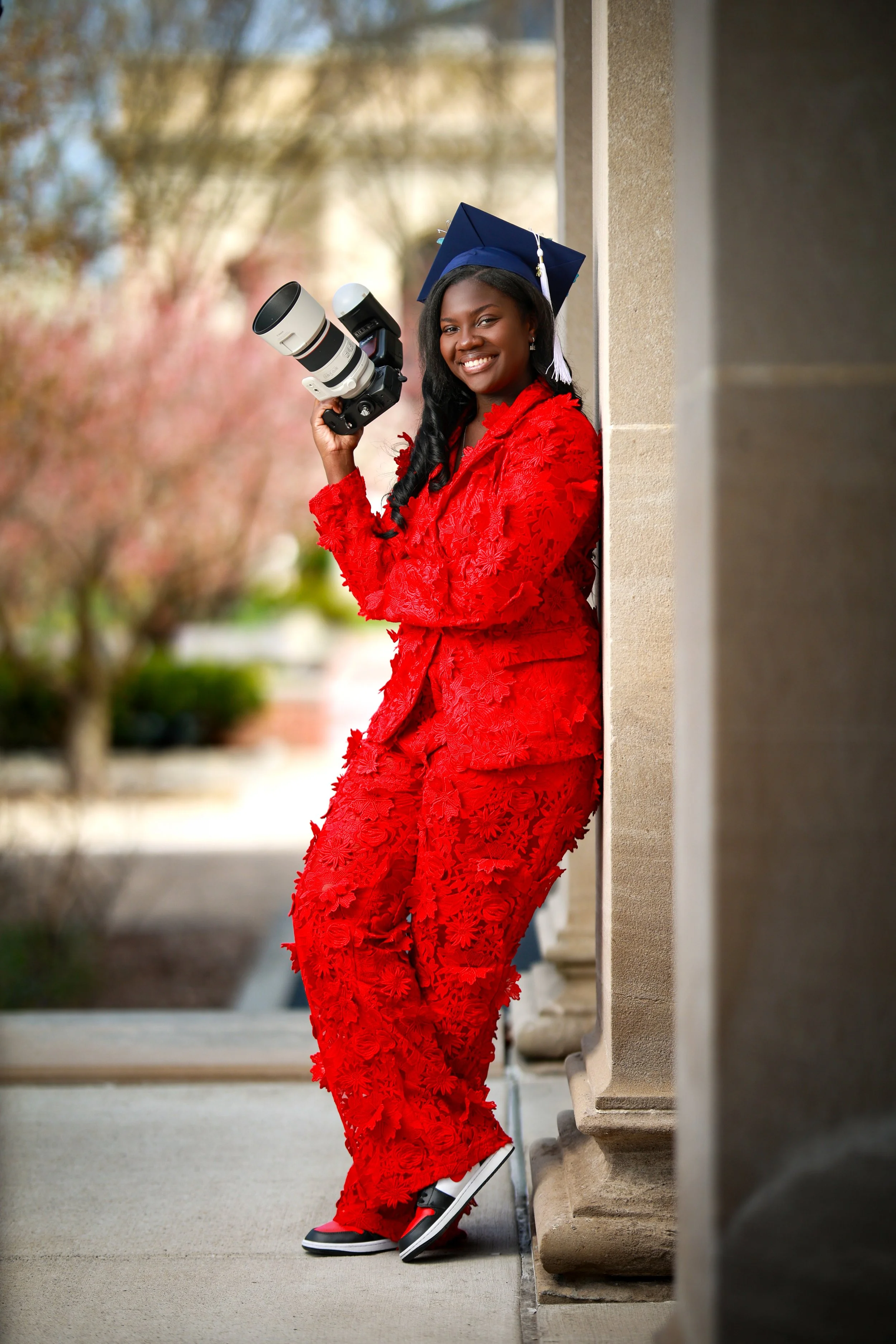 Young woman in a red floral dress and graduation cap leaning against a stone building with a camera in her hand, outdoors with trees in the background.