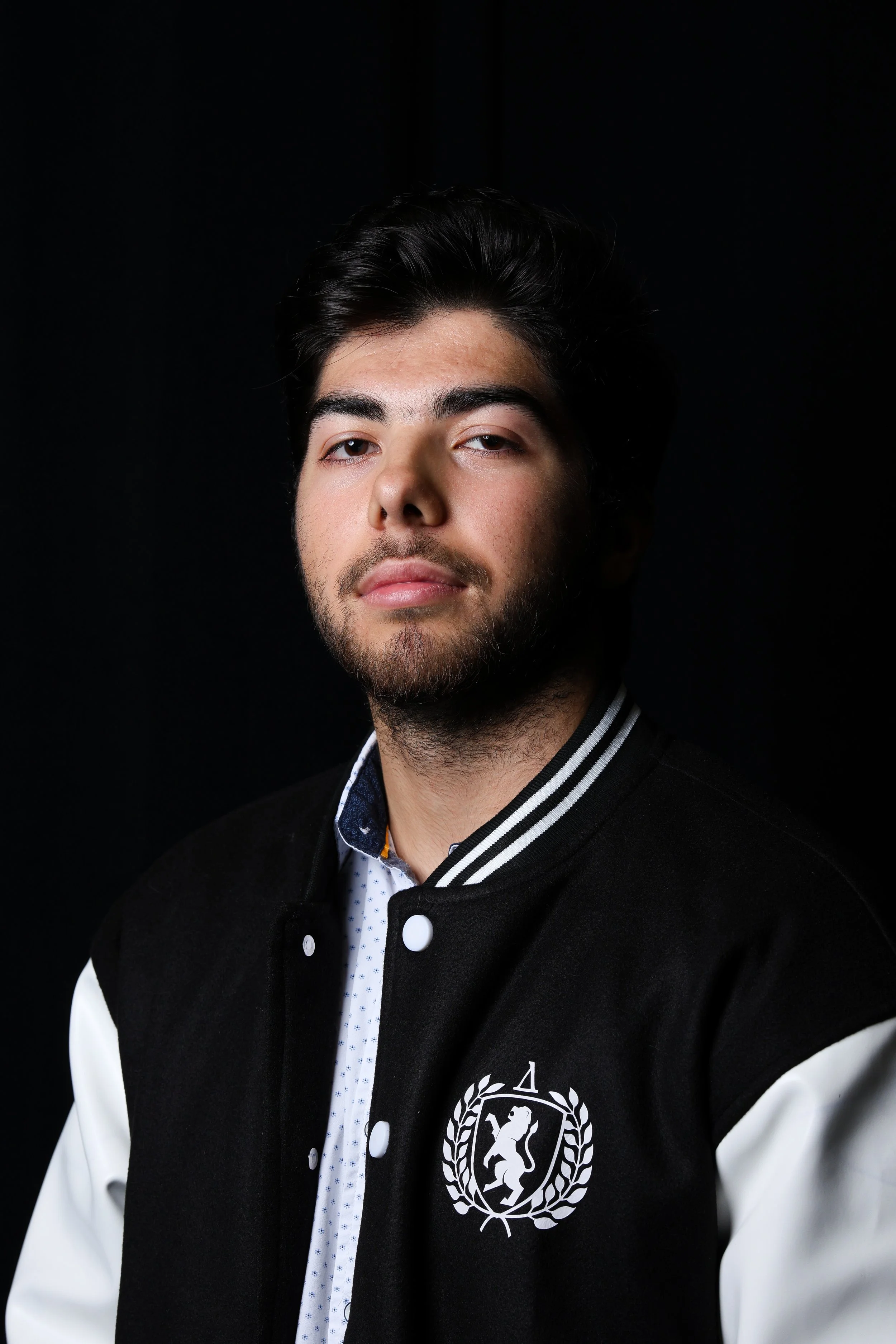 A young man with dark hair and light facial hair wearing a black and white varsity jacket with a crest emblem on the left side, standing against a black background.