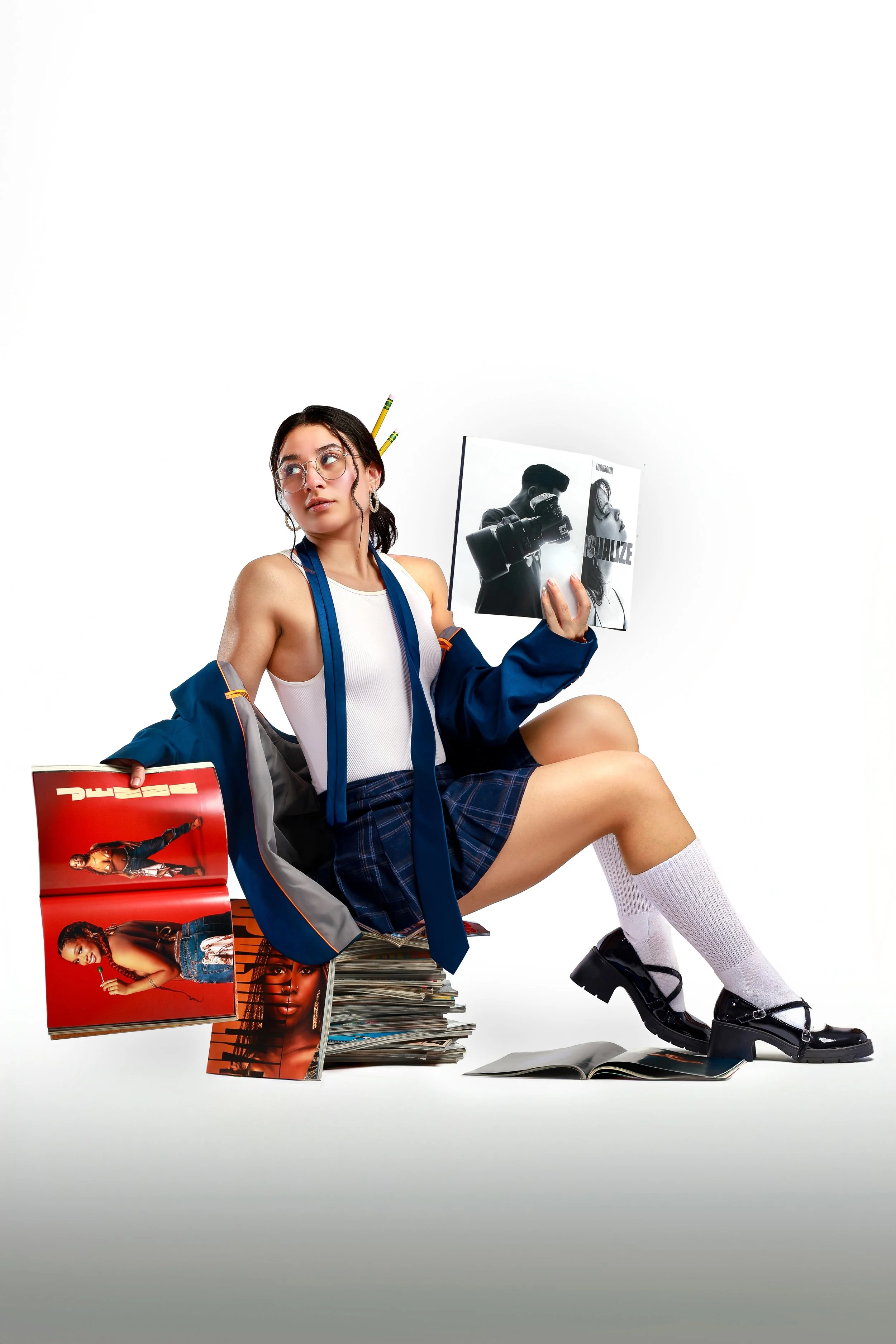Young woman in a school uniform holding a magazine, surrounded by other magazines and books, wearing glasses and accessories, sitting with one knee up against a white background.
