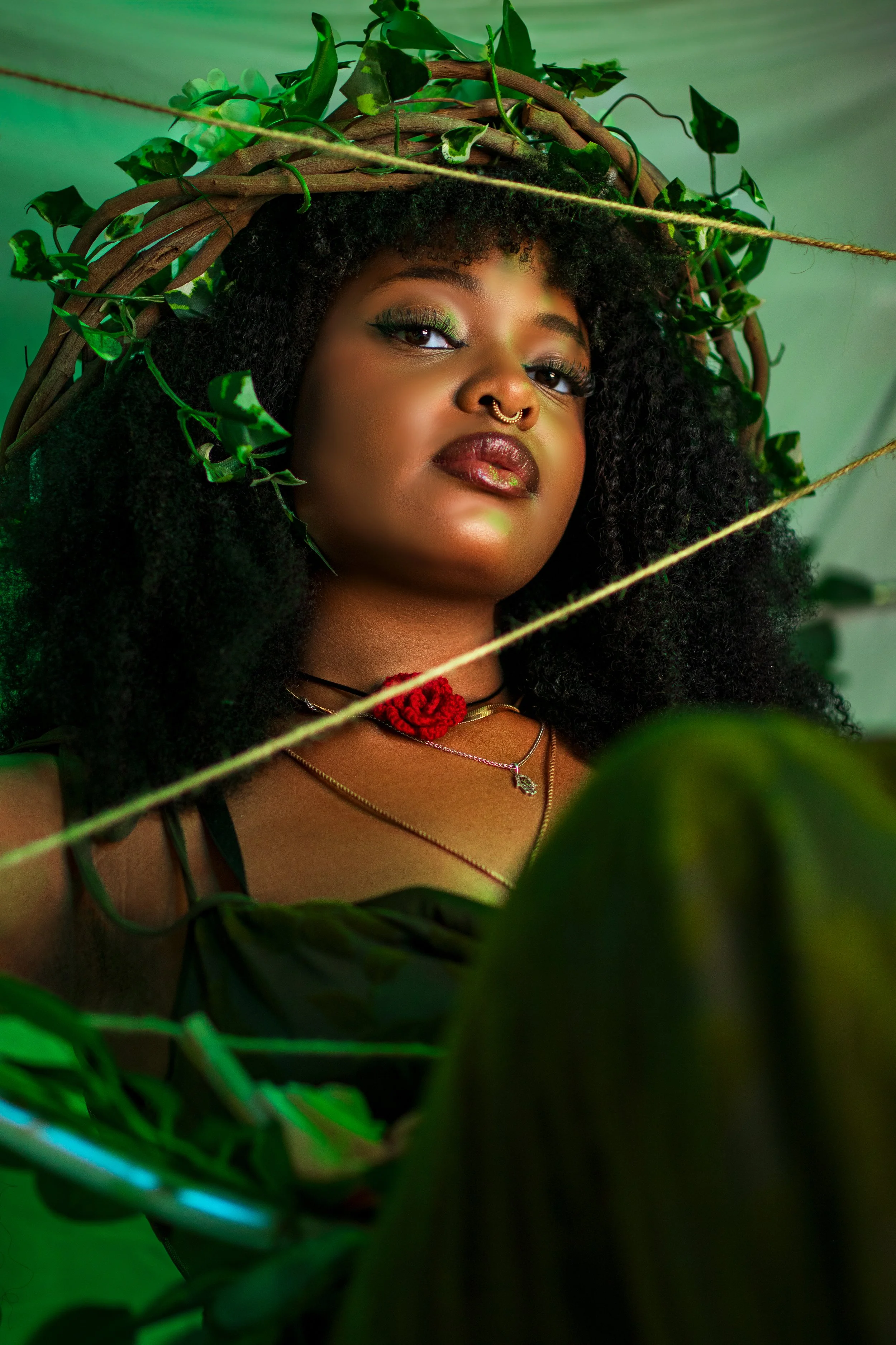 A woman with dark skin and curly black hair styled in a natural afro, wearing makeup with green eyeshadow and dark lipstick, adorned with a septum ring, jewelry including a necklace with a red flower, and surrounded by green leaves and vines, with a green background and a diagonal string or vine in the foreground.