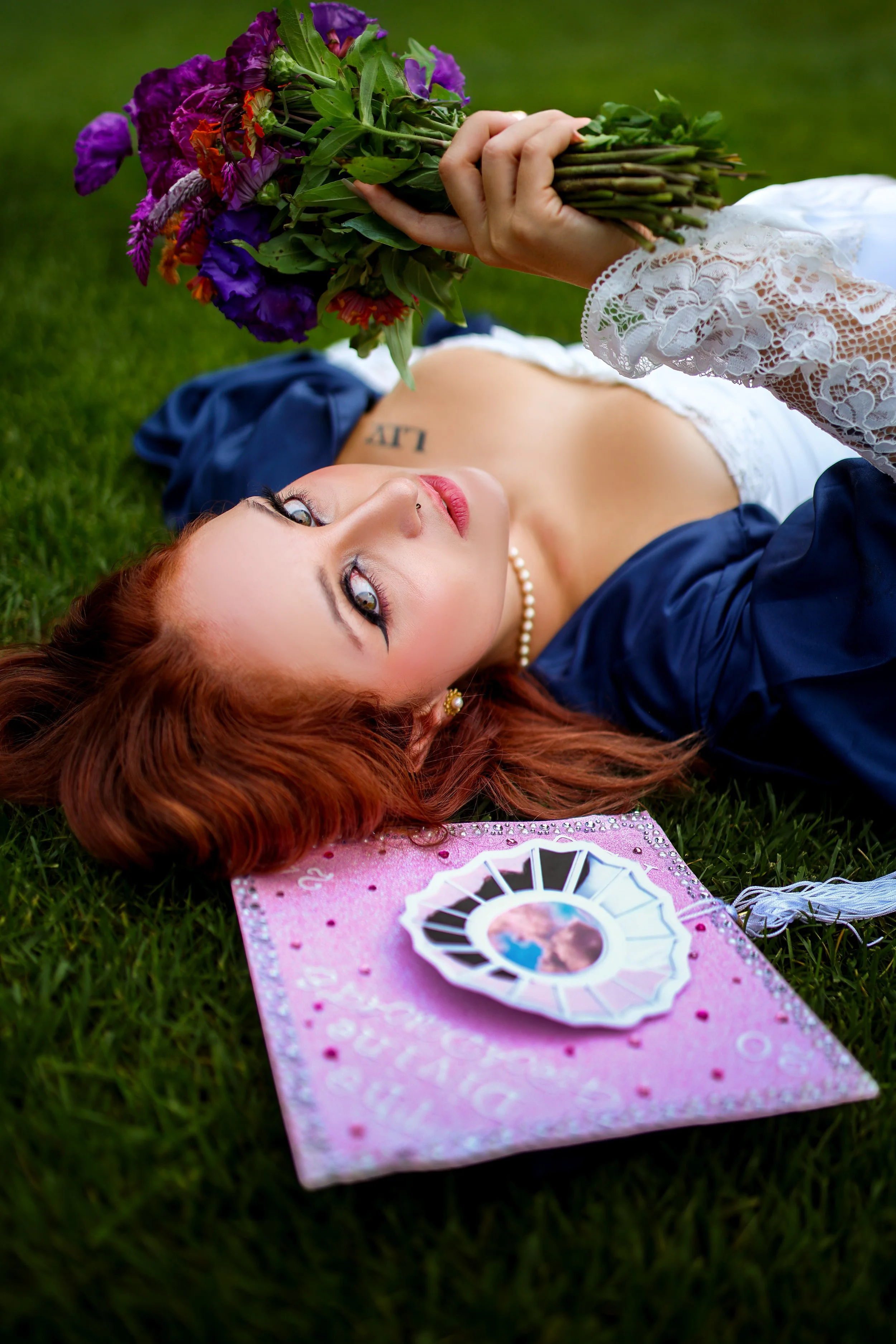 A woman with red hair lying on grass holding a bouquet of purple, orange, and green flowers. She is wearing a white lace top, pearl necklace, and blue jacket, with a tattoo visible on her chest. A pink dreamcatcher or decorative item is on the grass nearby.