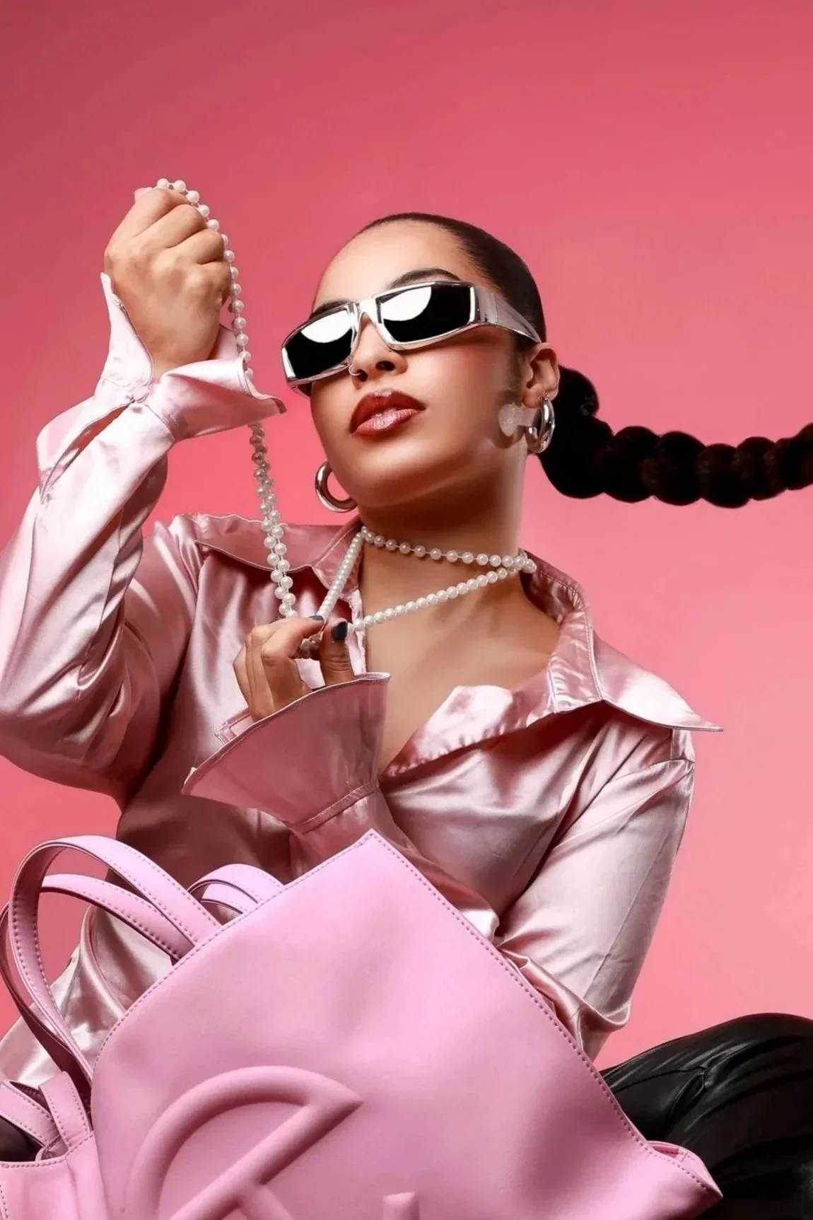 A woman in a shiny pink satin jacket, wearing large sunglasses, pearl jewelry, and earrings, holding a string of pearls in her hand against a pink background, with a pink handbag in front of her.