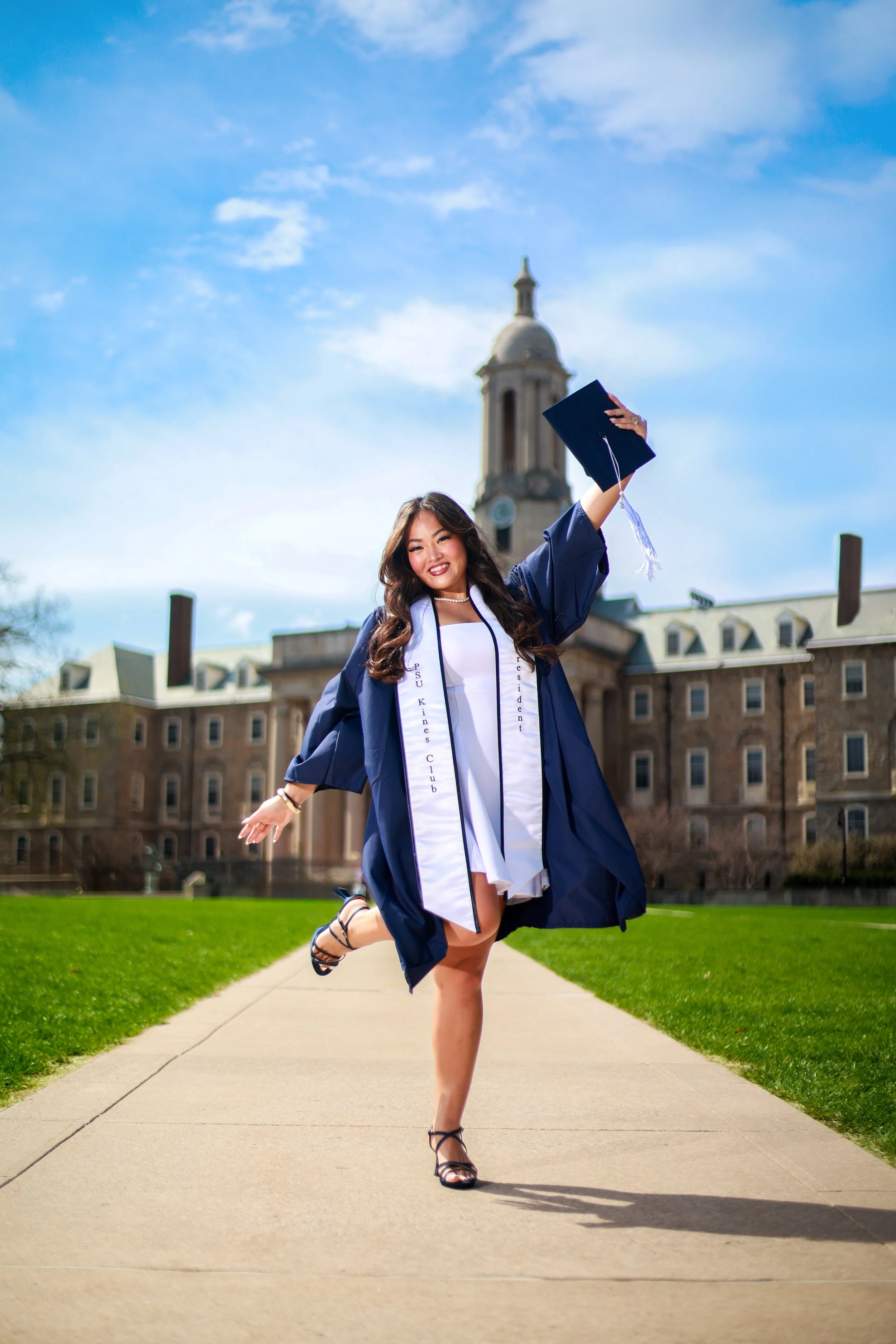 A woman in a graduation cap and gown celebrating on a college campus with a historic building and clock tower in the background.