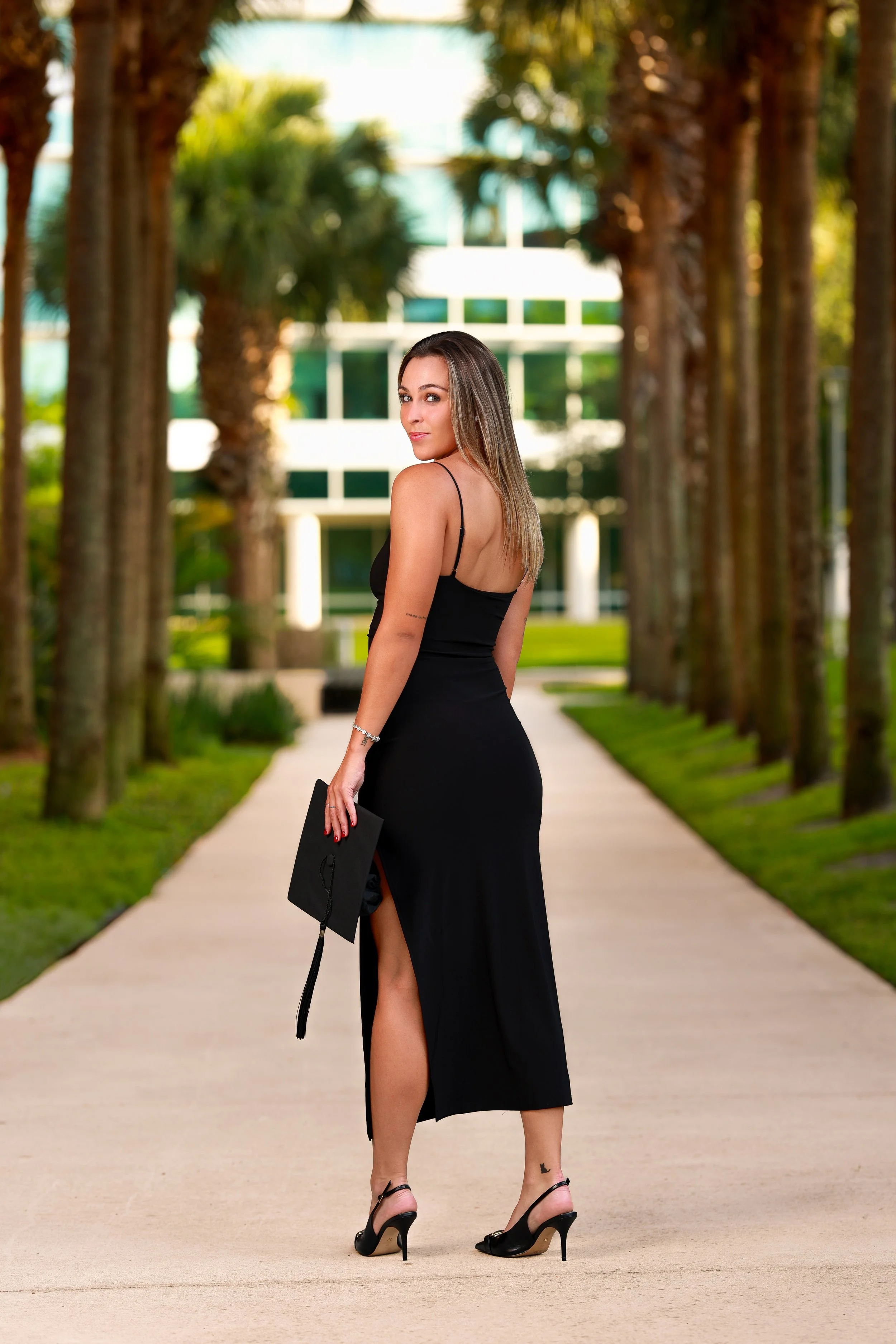 A woman in a black dress and high heels stands on a sidewalk holding a graduation cap with a cord. She is outdoors with tall trees lining the walkway and a building with green and white windows in the background.