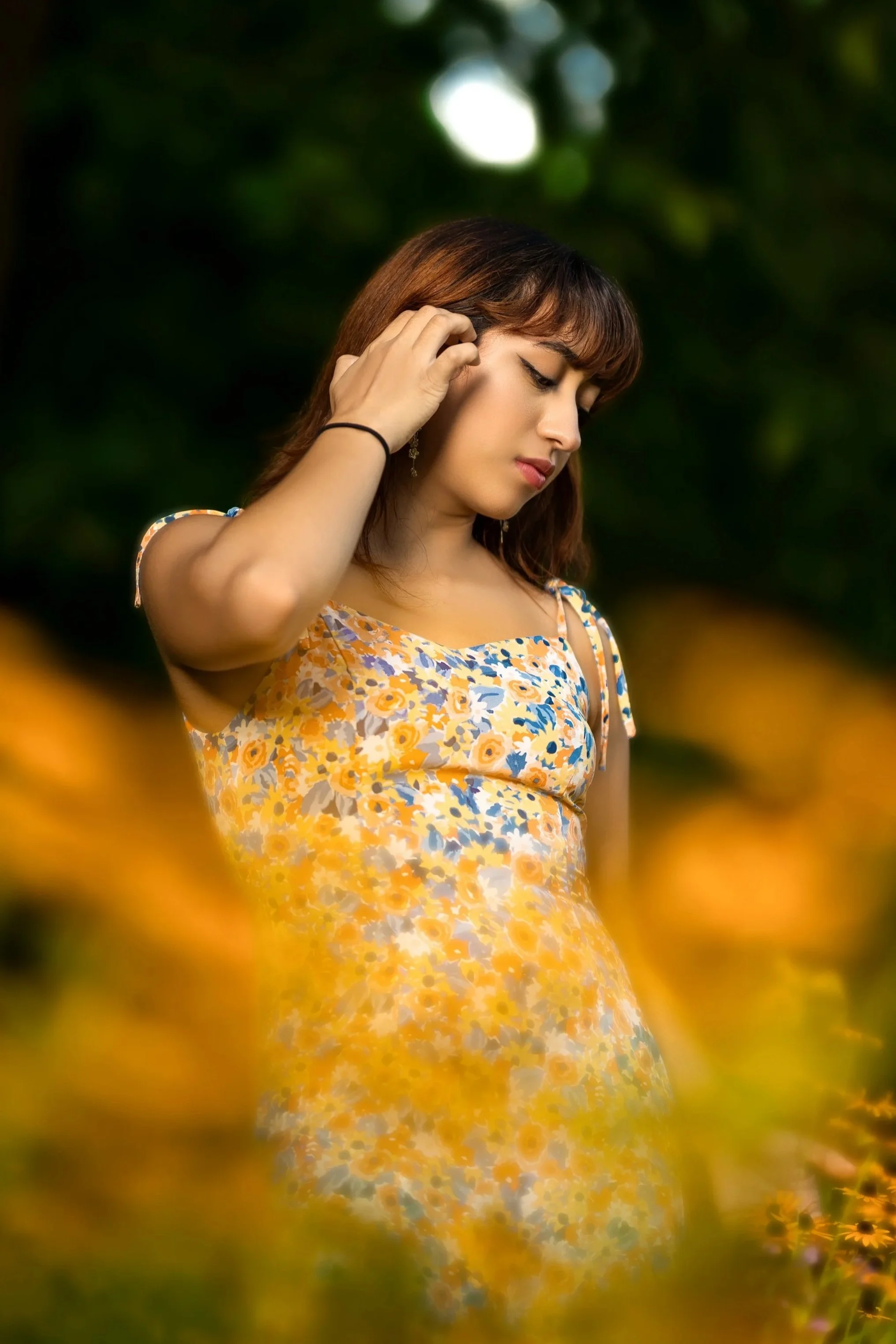 A young woman in a yellow flower-patterned dress with orange, yellow, and white flowers, standing outdoors among yellow and green foliage, with her eyes closed and her hand touching her ear.