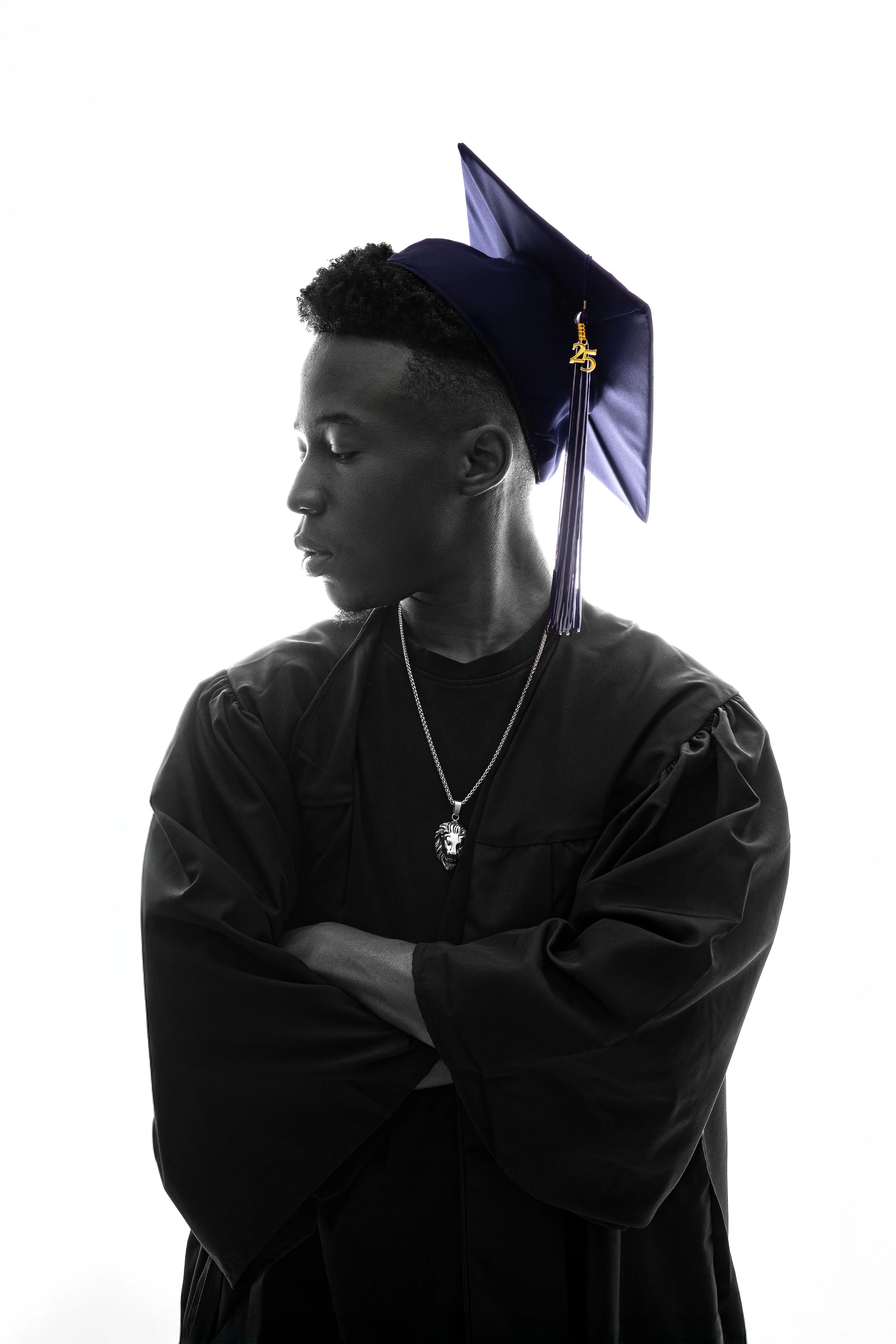 A young man with short curly hair, wearing a black jacket and jewelry, is posed with arms crossed against a plain white background. He is wearing a navy blue graduation cap with a gold '25' charm and tassel.