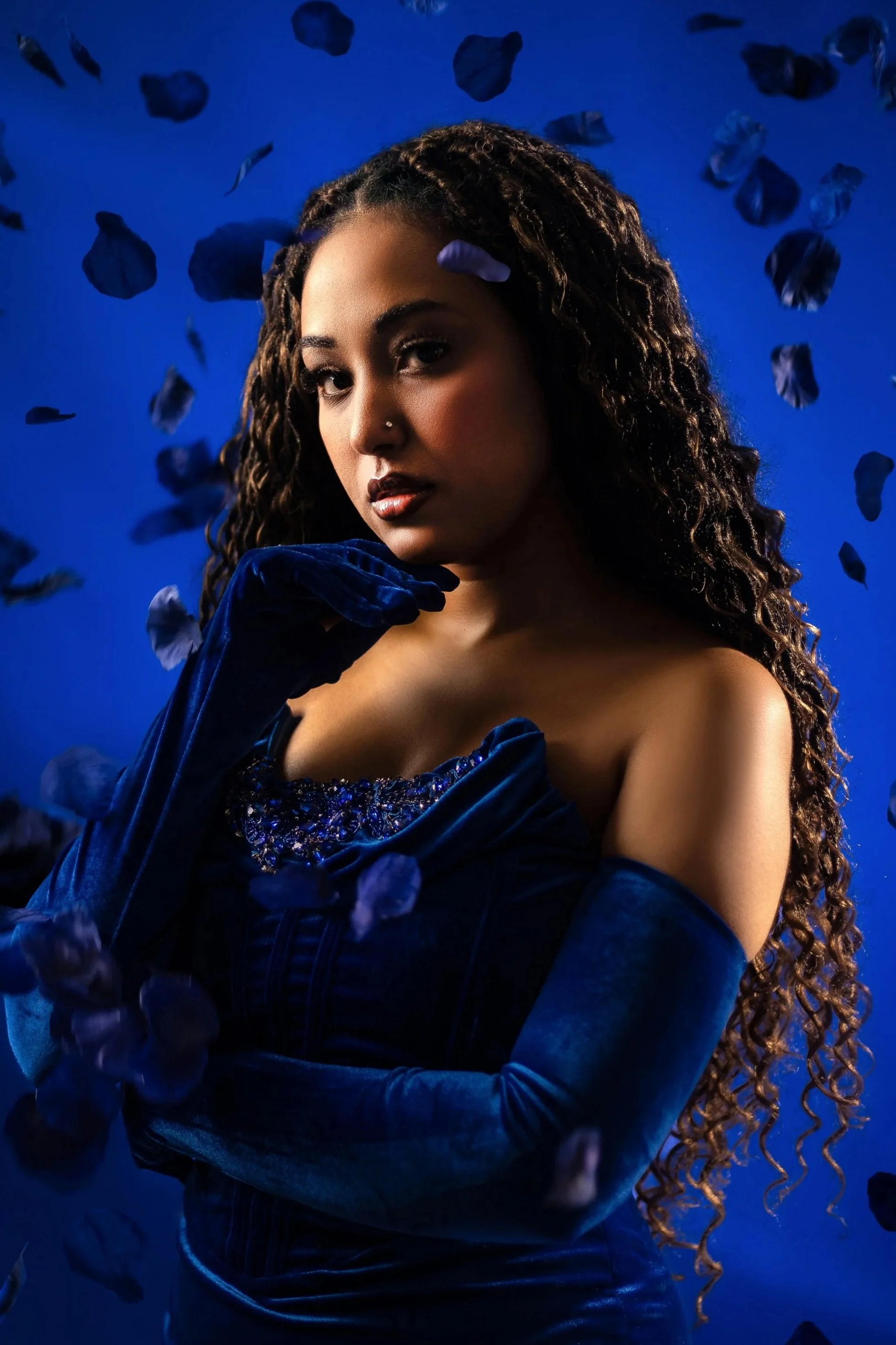 A woman with long, curly hair, dark lipstick, and a nose piercing, wearing dark blue elegant dress and gloves, posed against a deep blue background with floating dark petals or leaves.