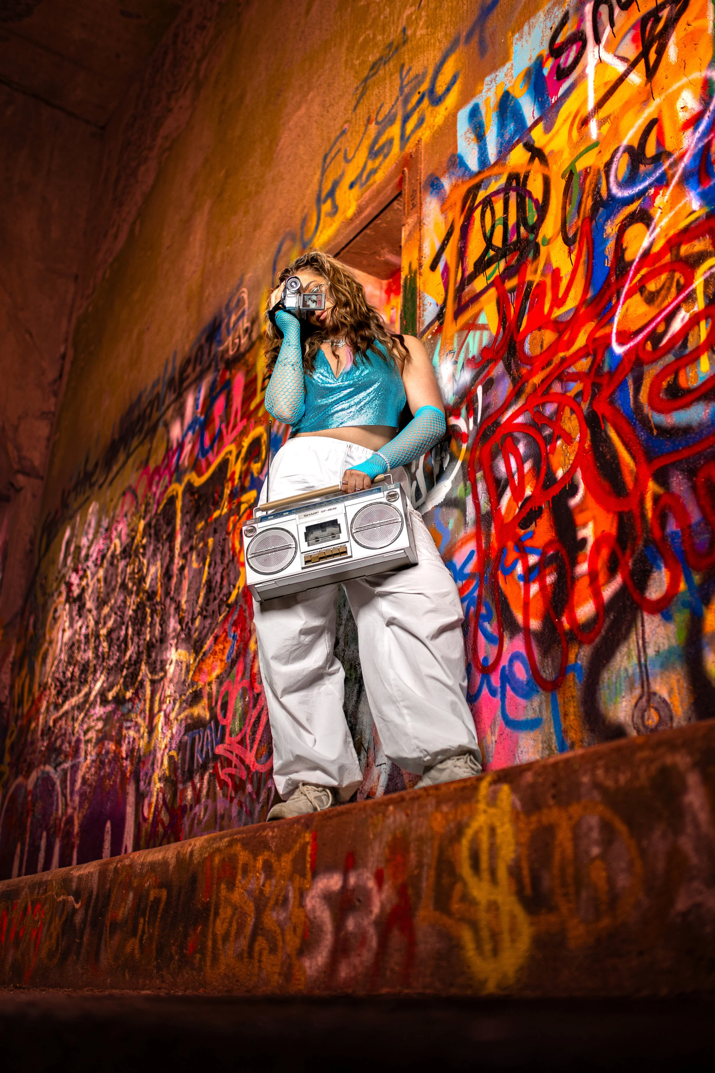 A woman in a teal top and white pants is standing in front of a graffiti-covered wall, holding a large boombox and pretending to take a picture with a vintage camera. The image is taken from a low angle, capturing colorful graffiti with vibrant orange, red, blue, and black writings.