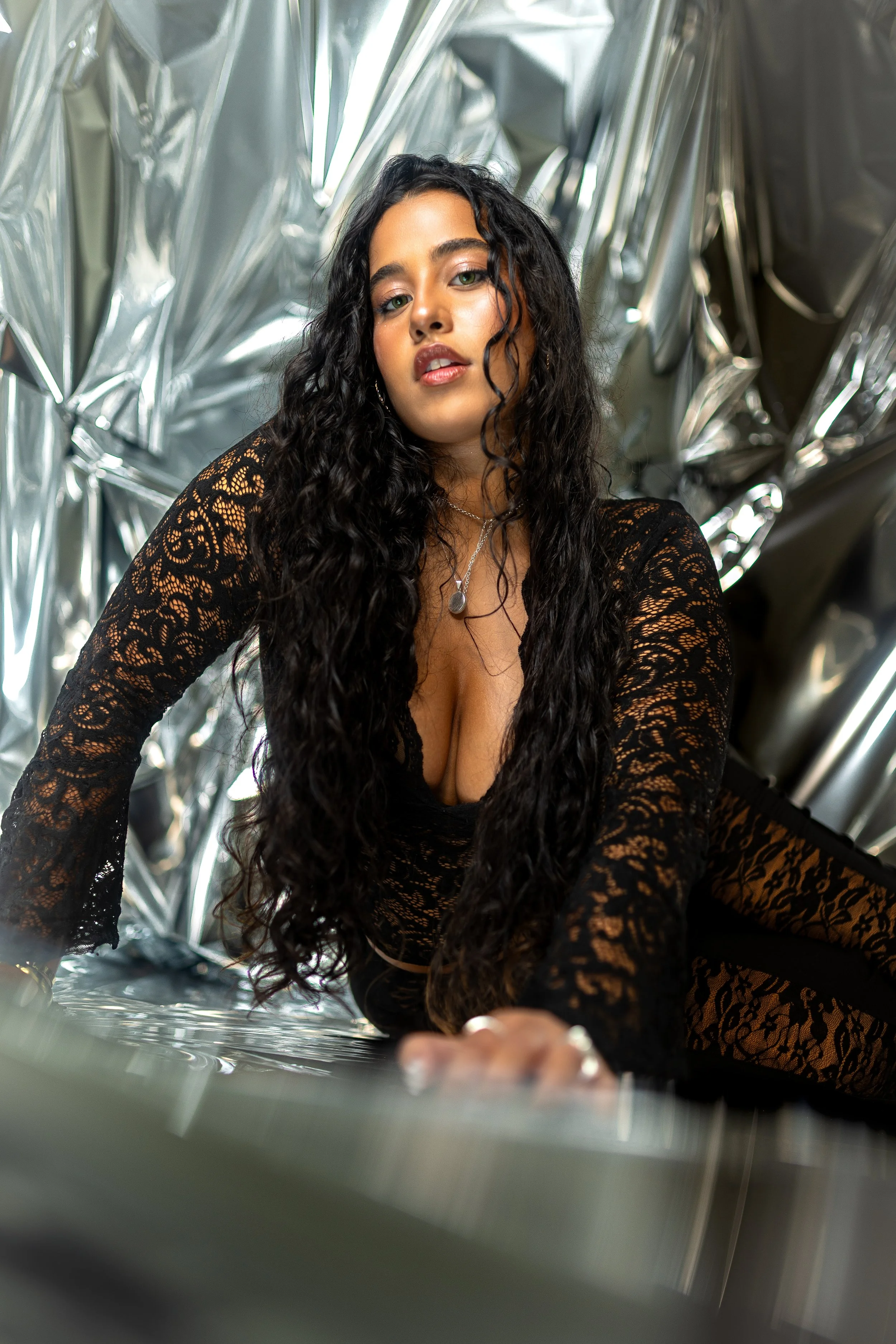 A woman with long, curly black hair wearing a black lace top and silver jewelry is posing for a photo in front of crumpled metallic foil background.