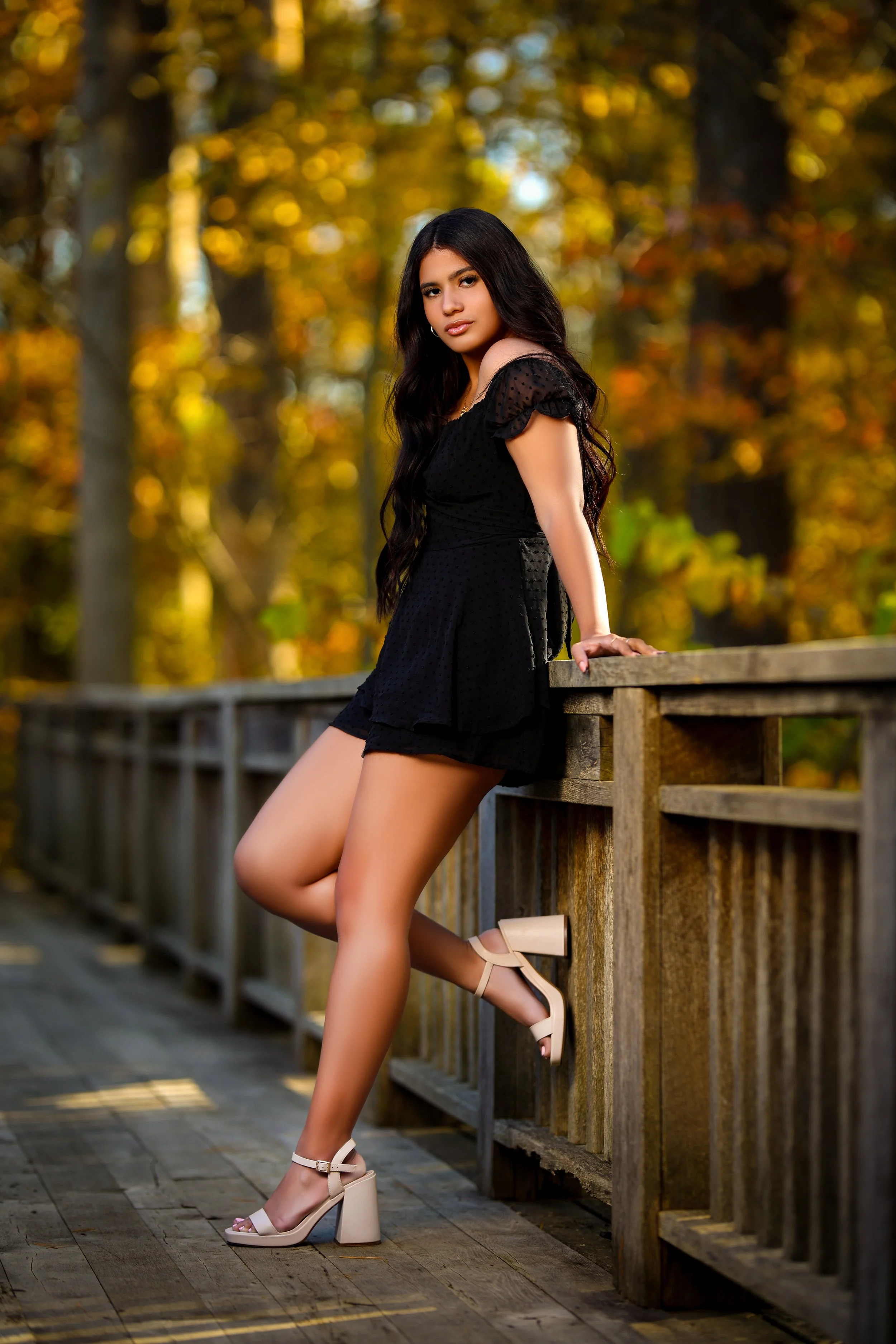 A young woman with long dark hair wearing a black off-shoulder dress and beige high-heeled sandals standing outdoors on a wooden pathway, leaning against a wooden railing with a background of blurred autumn trees.