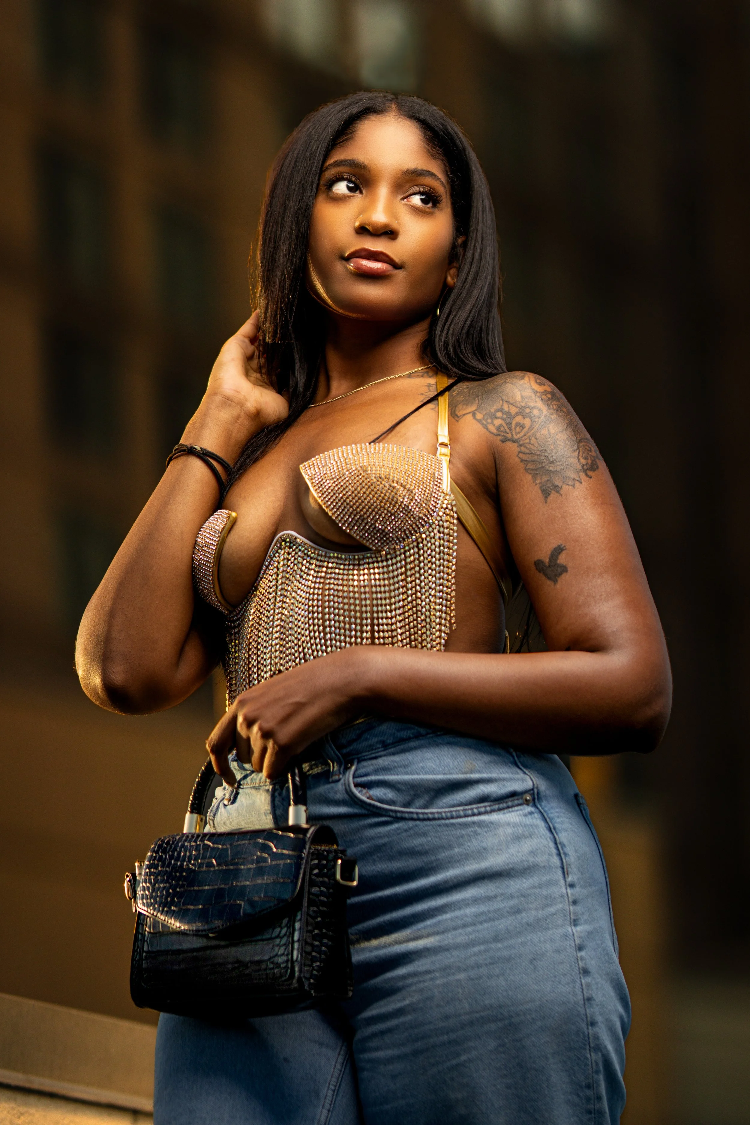 A young woman with dark hair, wearing a shiny gold crop top and blue jeans, standing in an indoor setting with a blurred background, holding a black purse with a textured pattern.