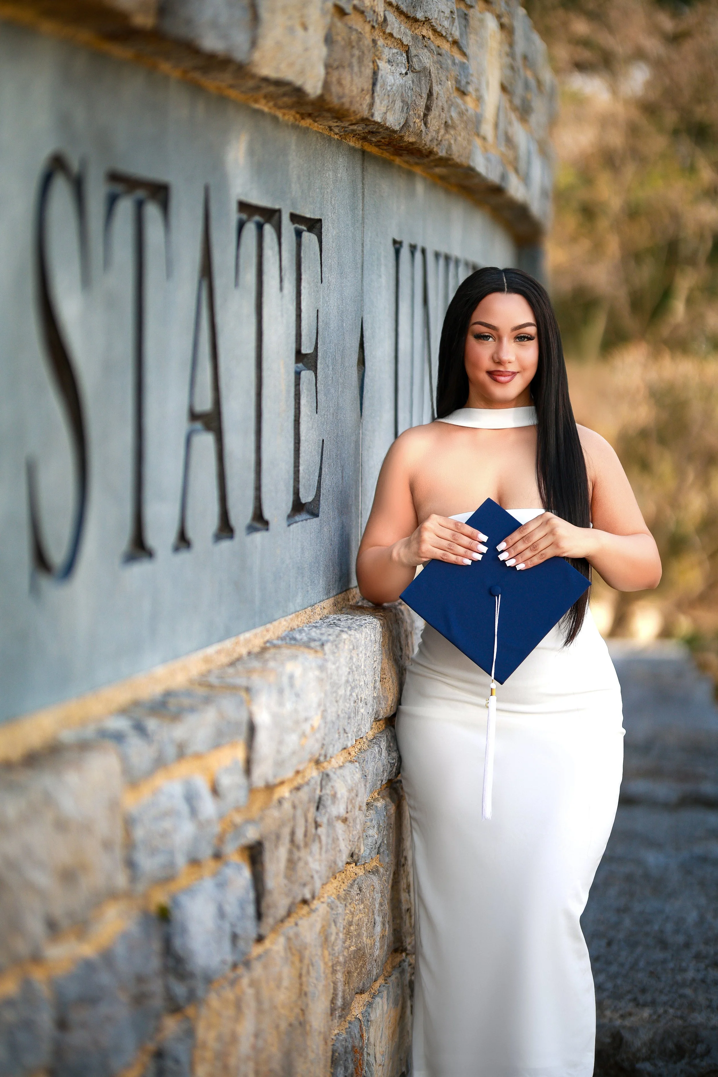 A young woman in a white dress holding a graduation cap in front of a stone wall with the writing 'CIVIC' on it.