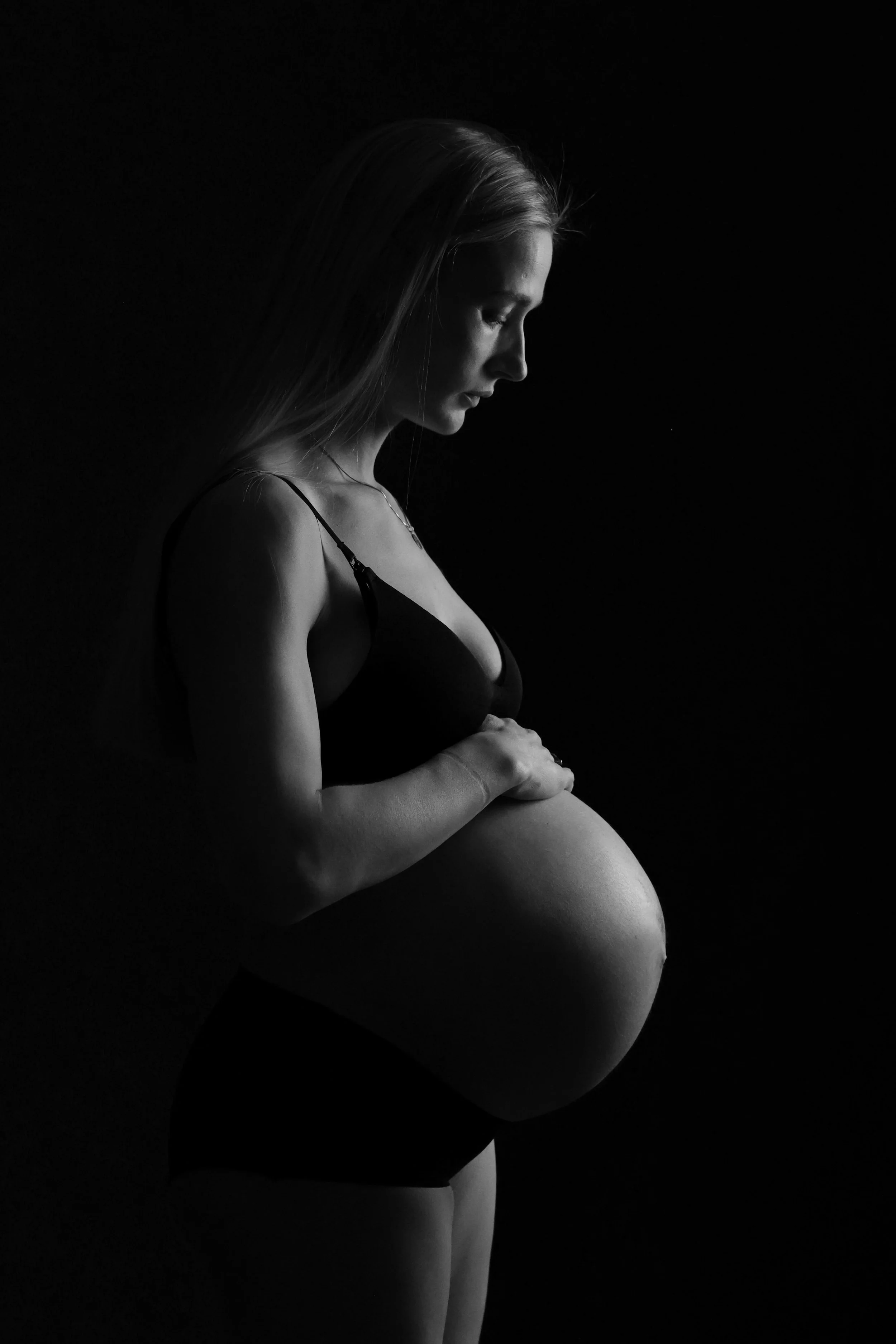 Svart-kvitt-bilete av ei gravid kvinne i profil som ser ned, med langt hår og eit minimalistisk antrekk mot svart bakgrunn