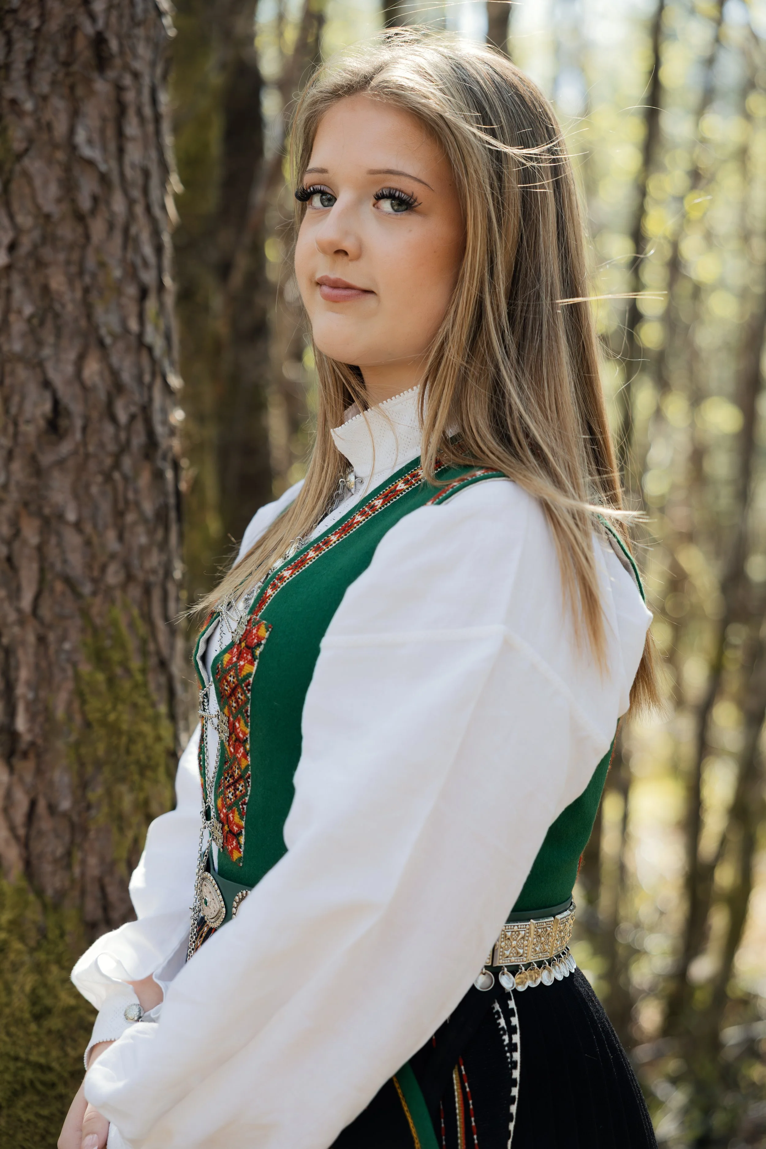 Ei ung kvinne i tradisjonell norsk bunad står ved eit tre i skogen, med sollys som skin gjennom trea.