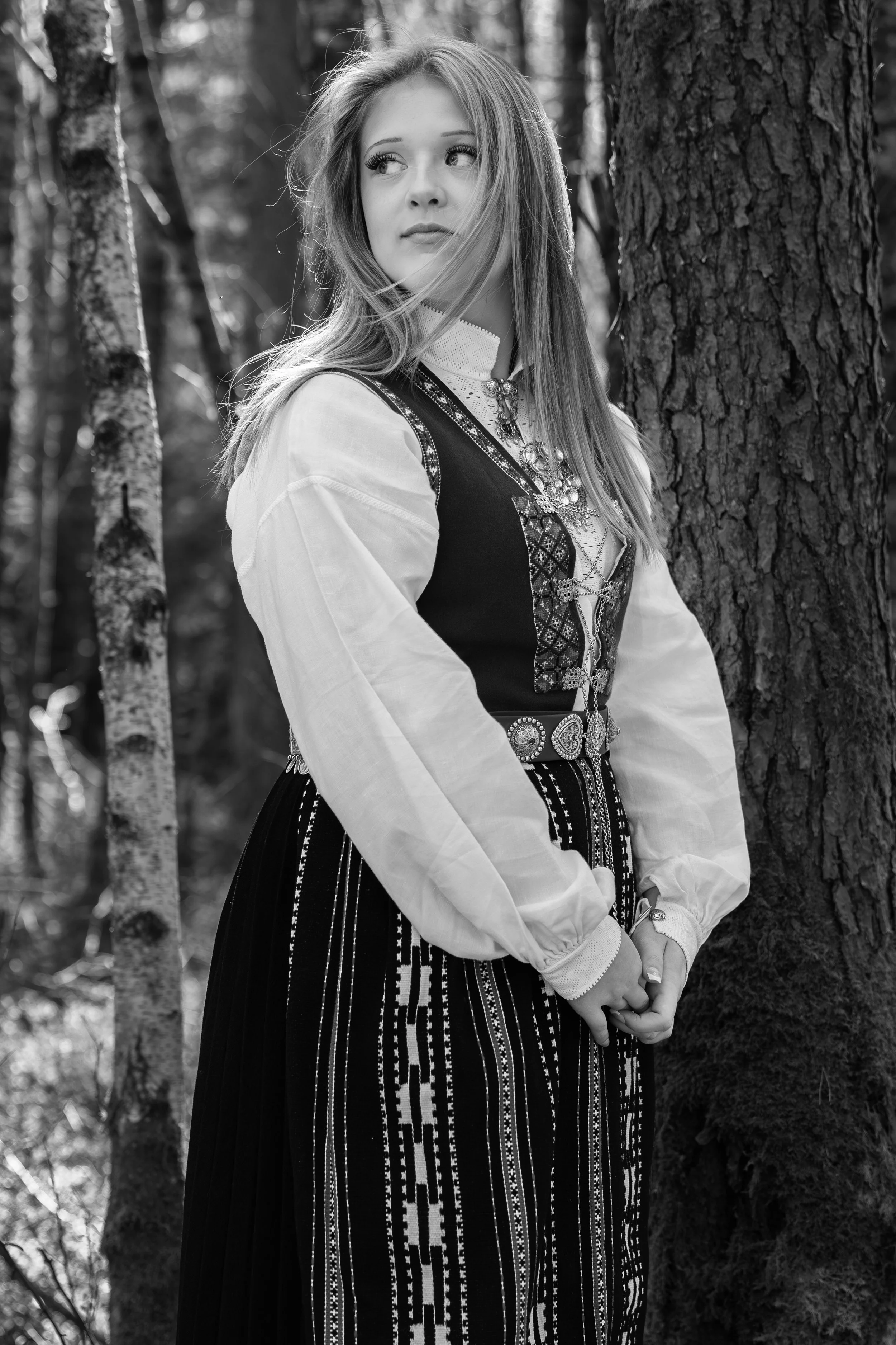 Svart-kvitt-bilete av ei ung kvinne i tradisjonell norsk bunad, ståande i ein skog omgitt av tre.