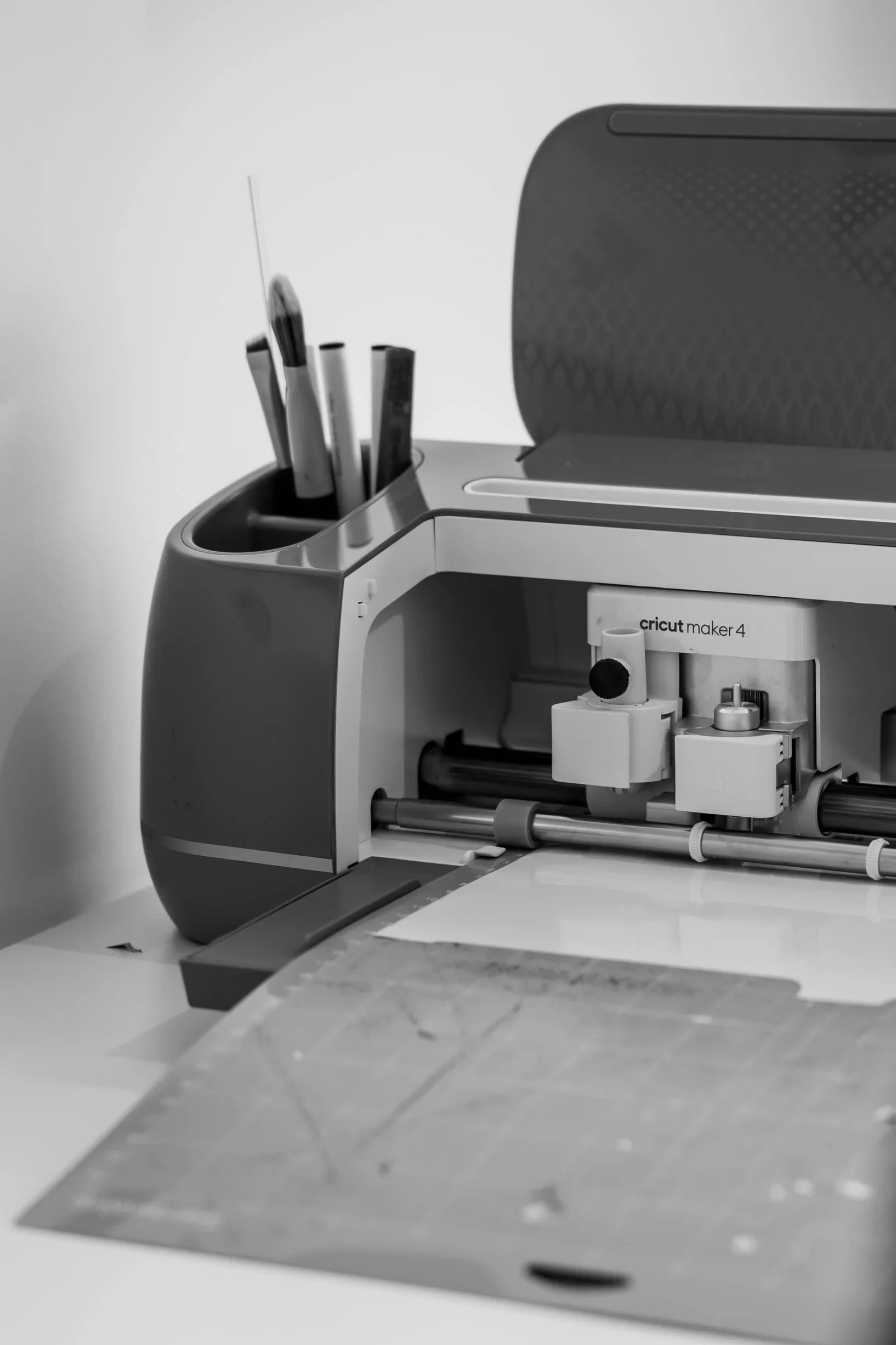 Svart-kvitt-bilete av ein Cricut Maker 4-kuttemaskin med ope lokk og pennar i oppbevaringsrommet, plassert på eit bord.