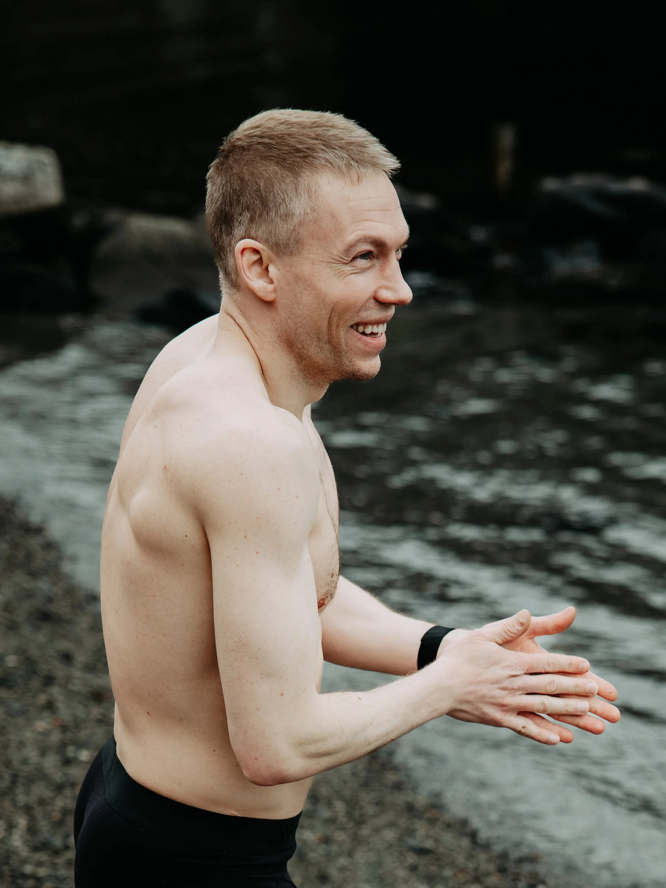 Ein mann med kort, lyst hår ler og står på ein steinstrand ved ein elv eller sjølv, og ser mot høgre. Han er uten skjorte og har på seg svarte treningsbukser og eit armband.