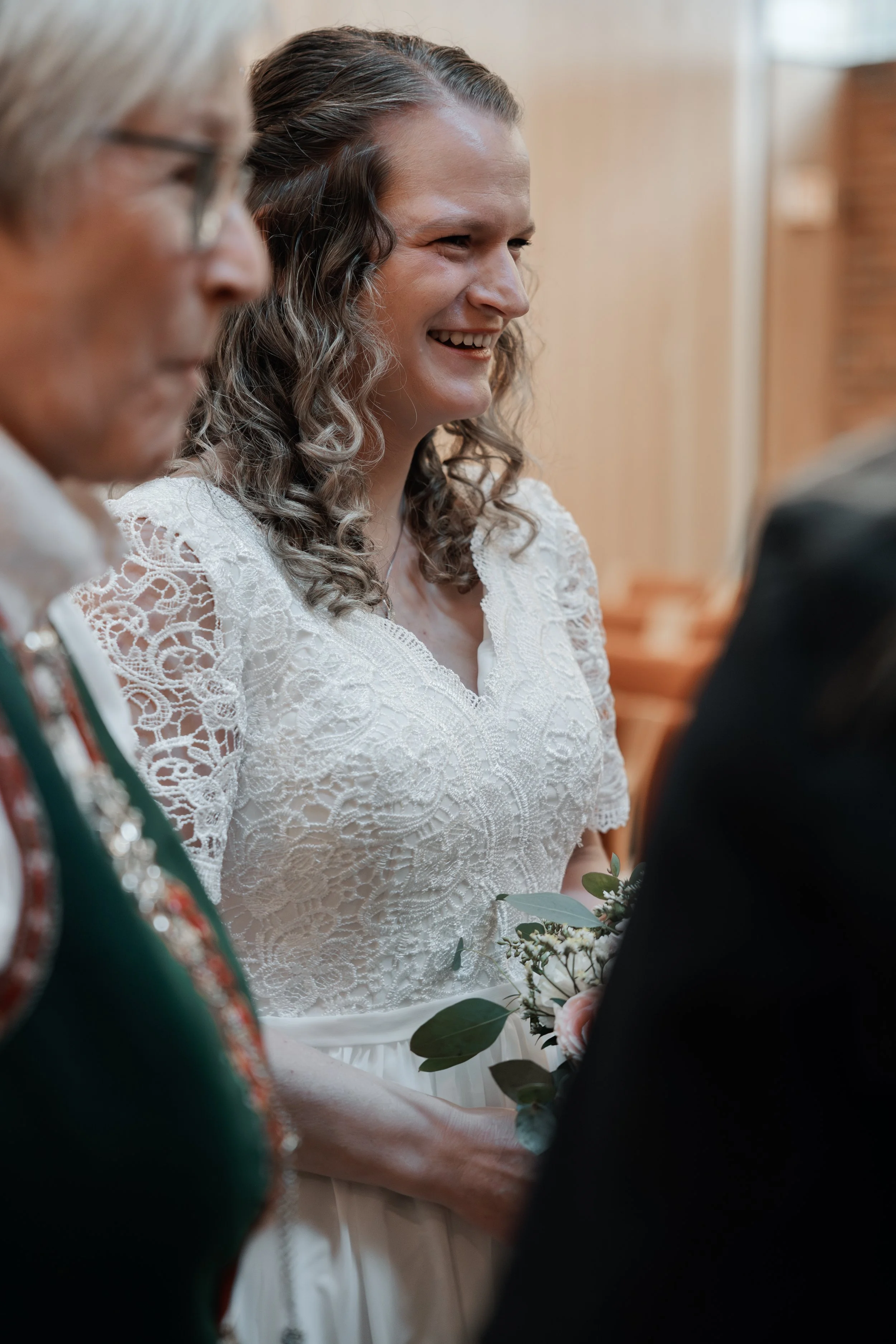Ei brud med langt, krølla hår som smiler under vigsla, iført ei kvit brudekjole med blonder og omgitt av gjester i formelle klede.