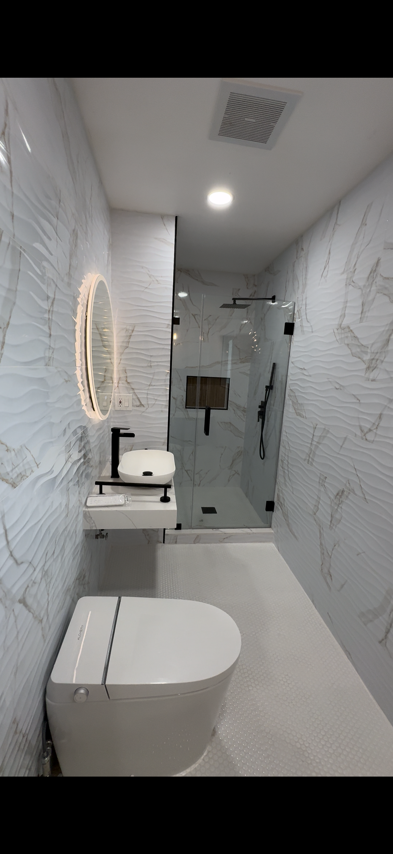 Modern bathroom with white textured marble walls, a wall-mounted sink with a black faucet, a round illuminated mirror, a toilet, and a walk-in shower with a glass door and black fixtures.