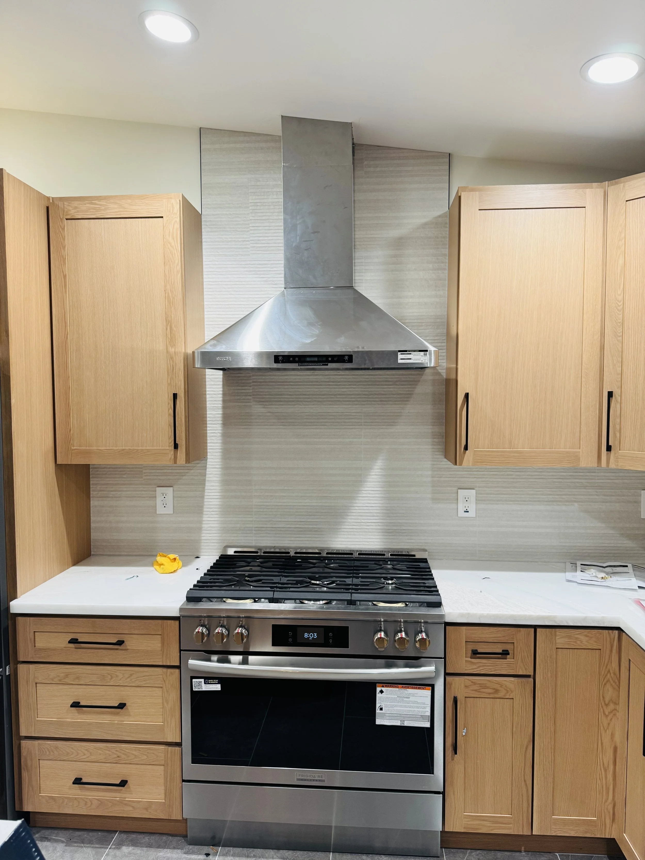 Kitchen with wooden cabinets, stainless steel range hood and stove, light-colored countertop, wall outlet, and some paper and a yellow object on the counter.