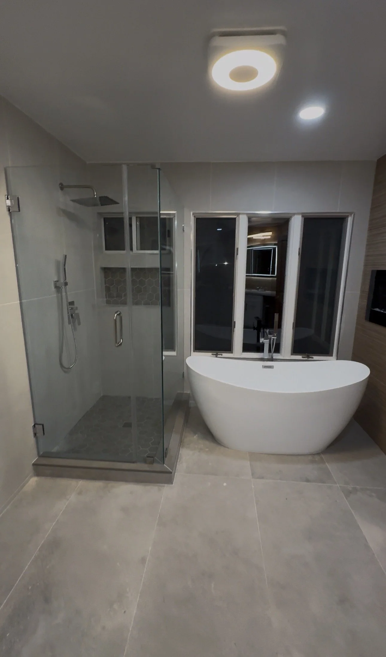 Modern bathroom with walk-in shower, bathtub, gray tile flooring, and windows.