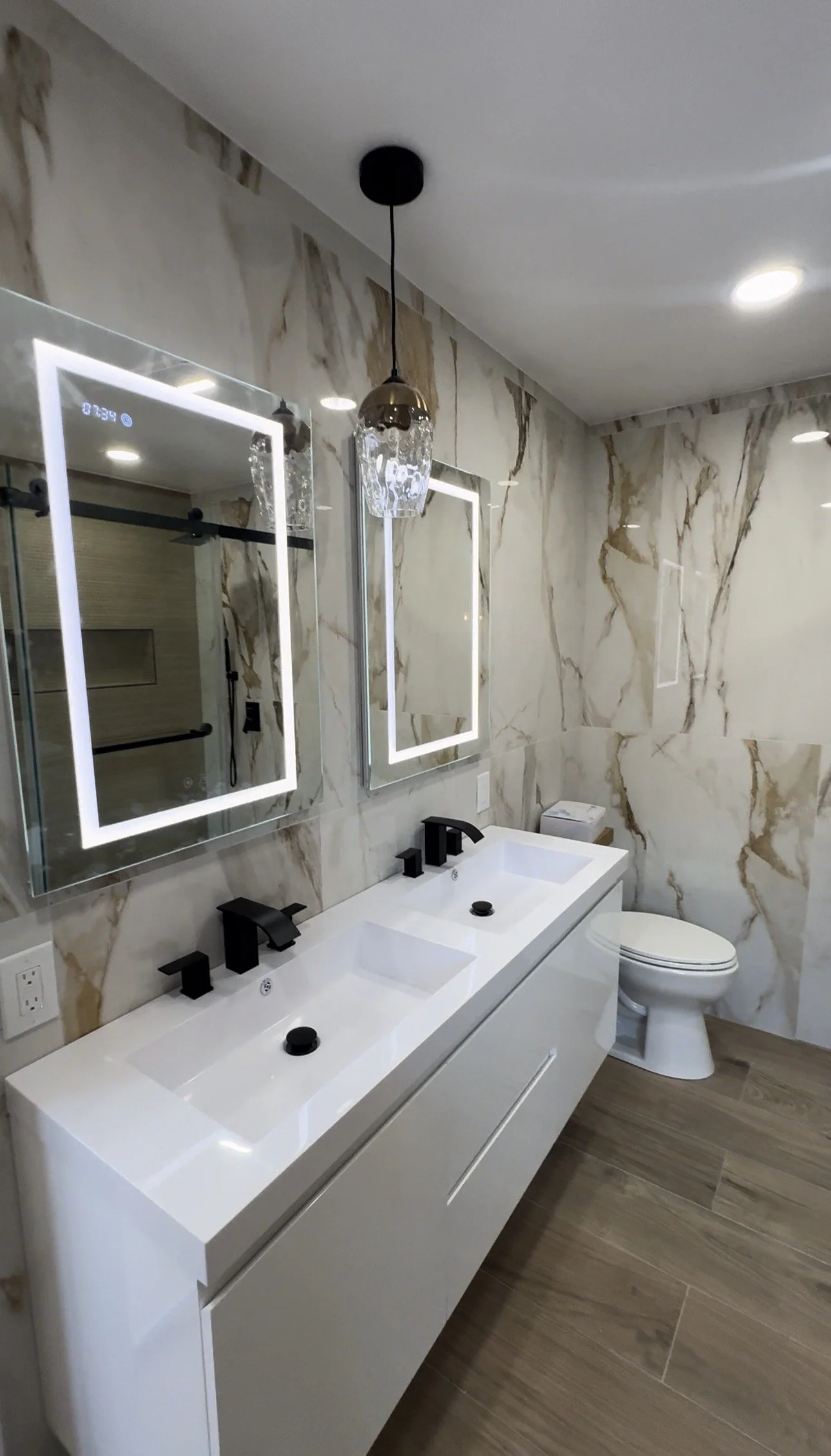 Modern bathroom with double sinks, black fixtures, illuminated mirrors, marble wall, pendant light, and toilet.
