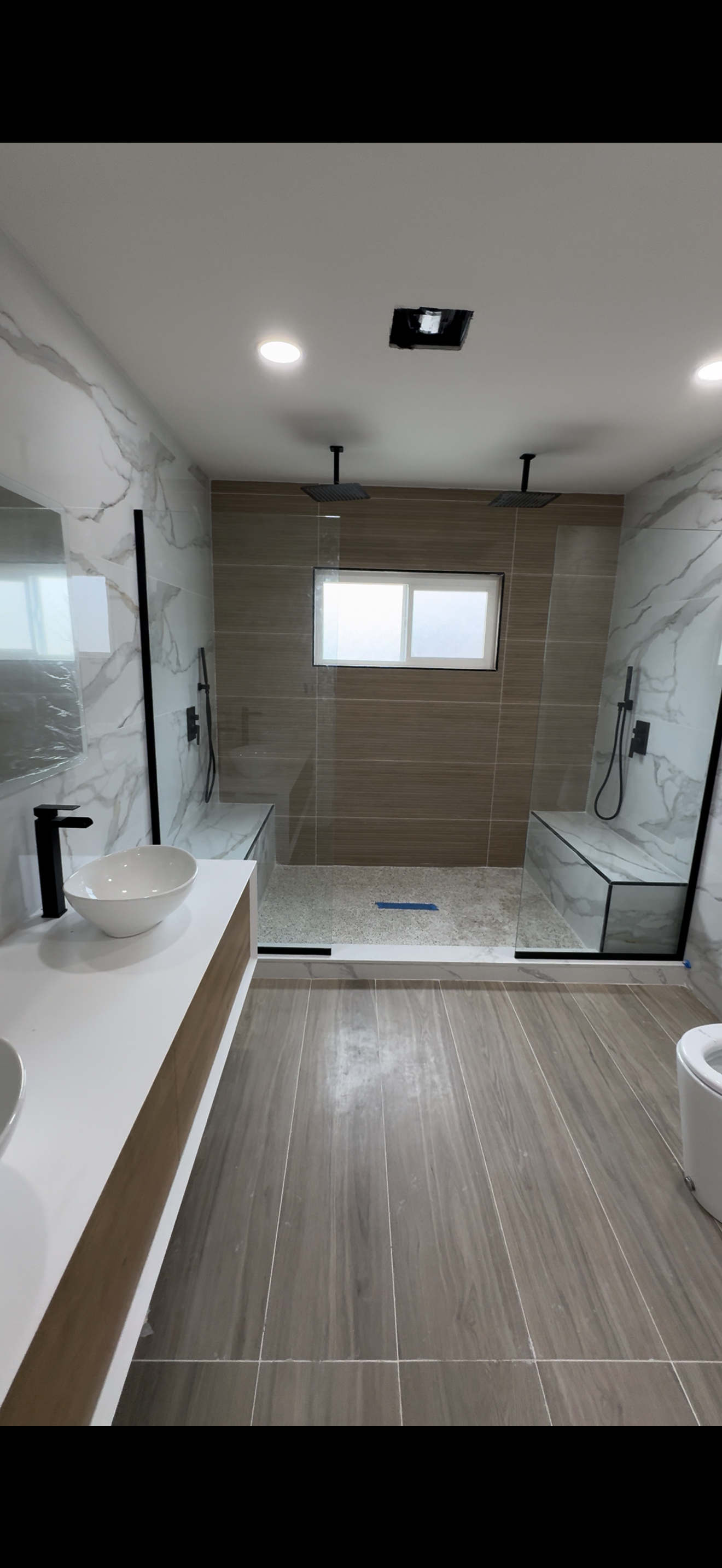 Modern bathroom with a walk-in shower separated by a glass partition, featuring dual rainfall showerheads, beige and marble tiles, a small frosted window, a vessel sink on a wooden vanity, and wood-look floor tiles.