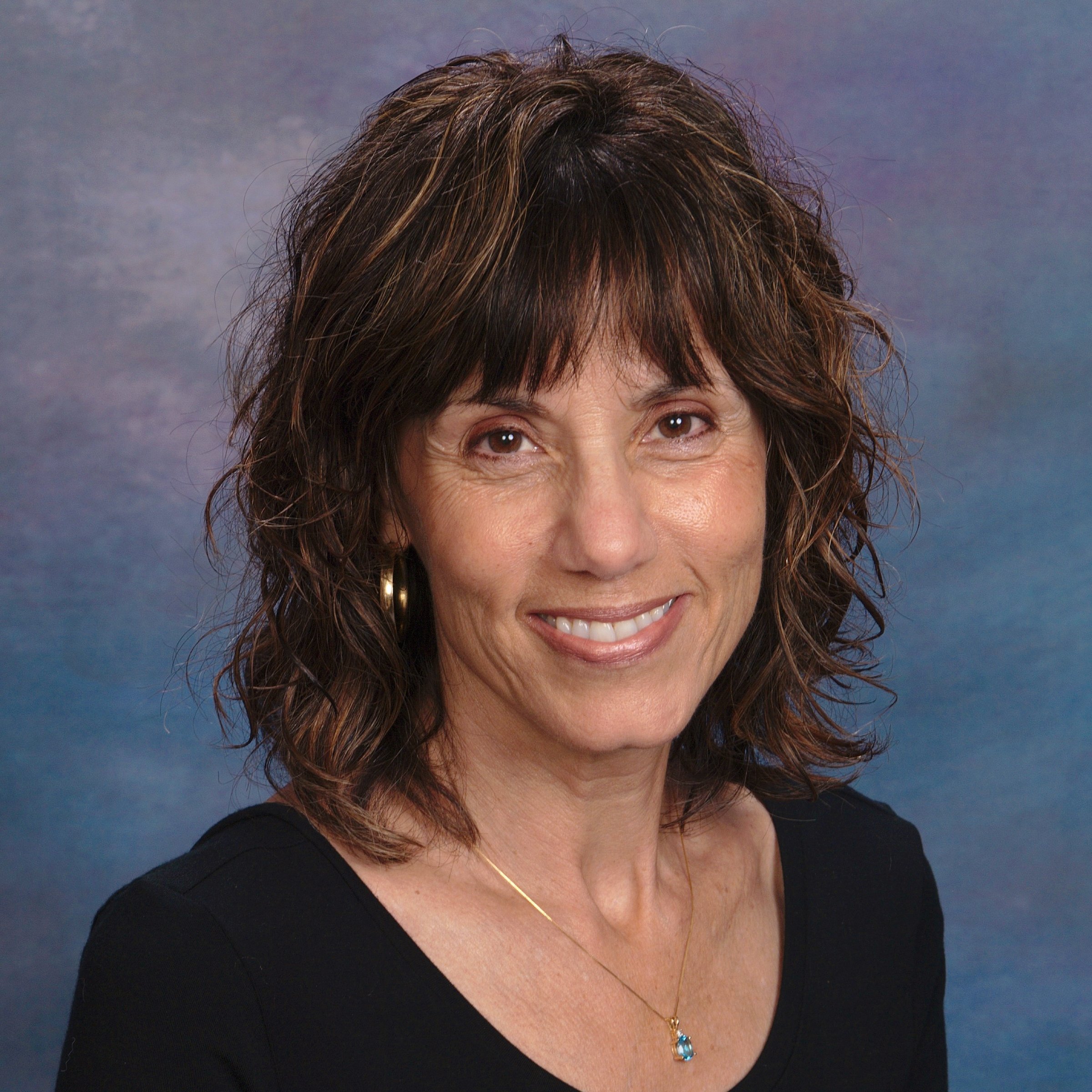 A woman with curly brown hair and a smile, wearing a black top, gold earrings, and a gold necklace with a blue pendant, against a cloudy blue background.