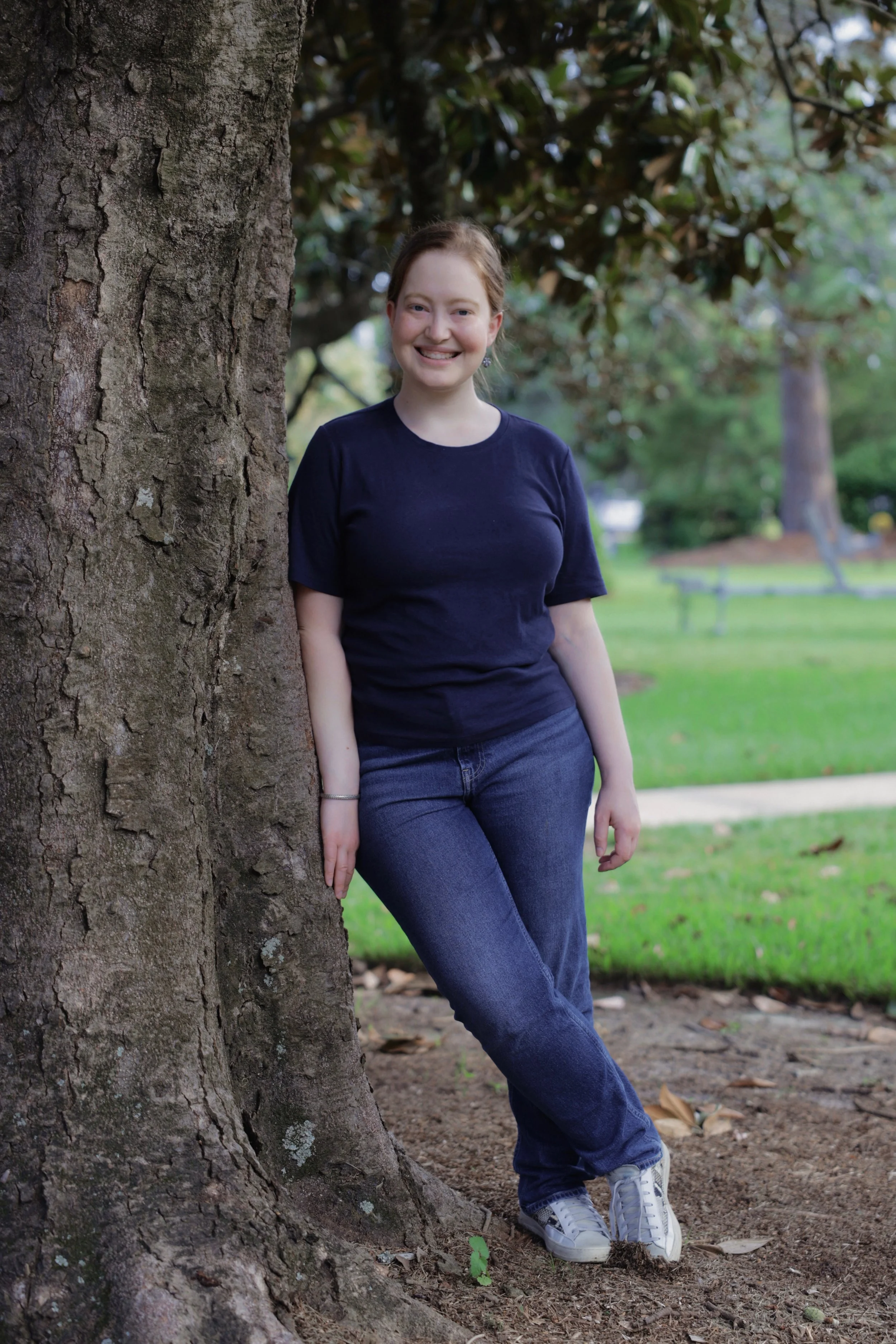 Lillie leans against a large magnolia tree and smiles at the camera. She is wearing a navy blue t shirt and blue jeans with gold and white sneakers.