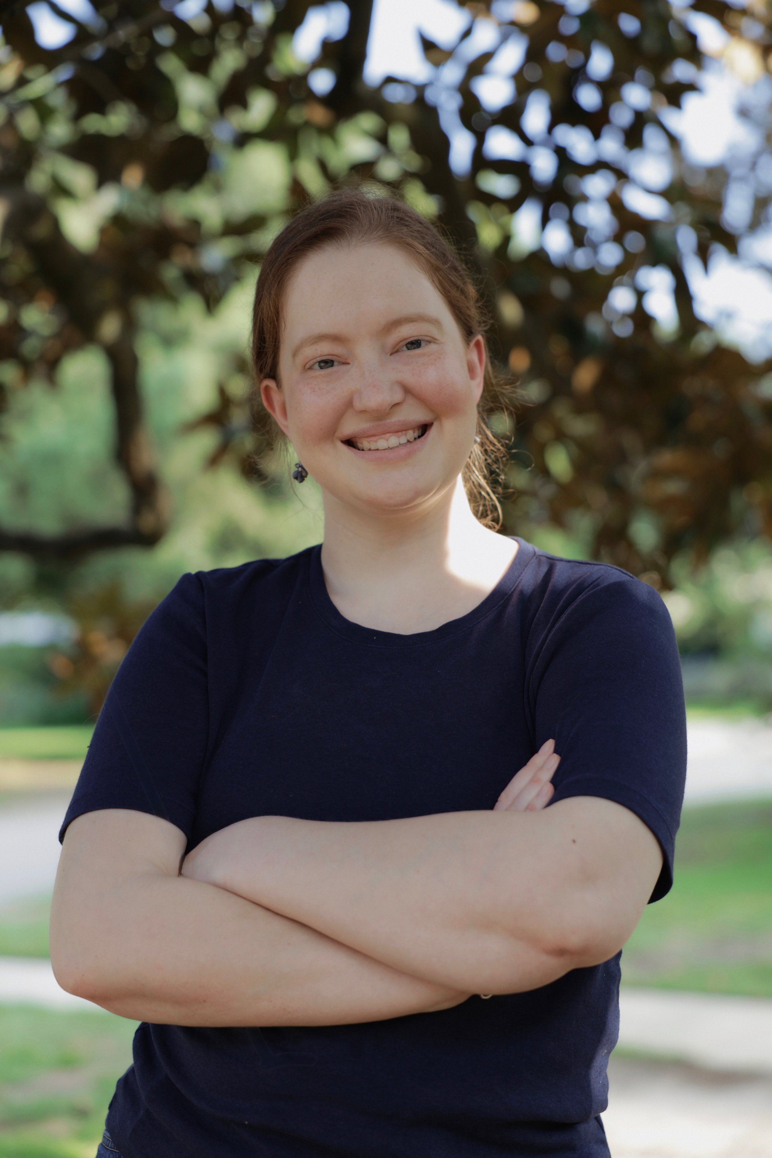 Lillie smiles at the camera with her arms crossed. She is wearing a navy blue t shirt and standing in front of a large magnolia tree.