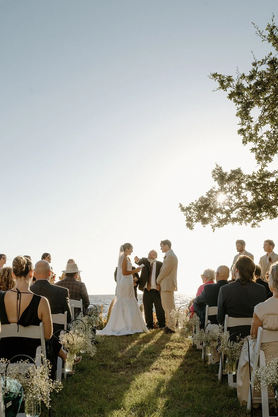 A wedding ceremony taking place outdoors by the beach during sunset with the bride and groom standing with an officiant, surrounded by seated guests.