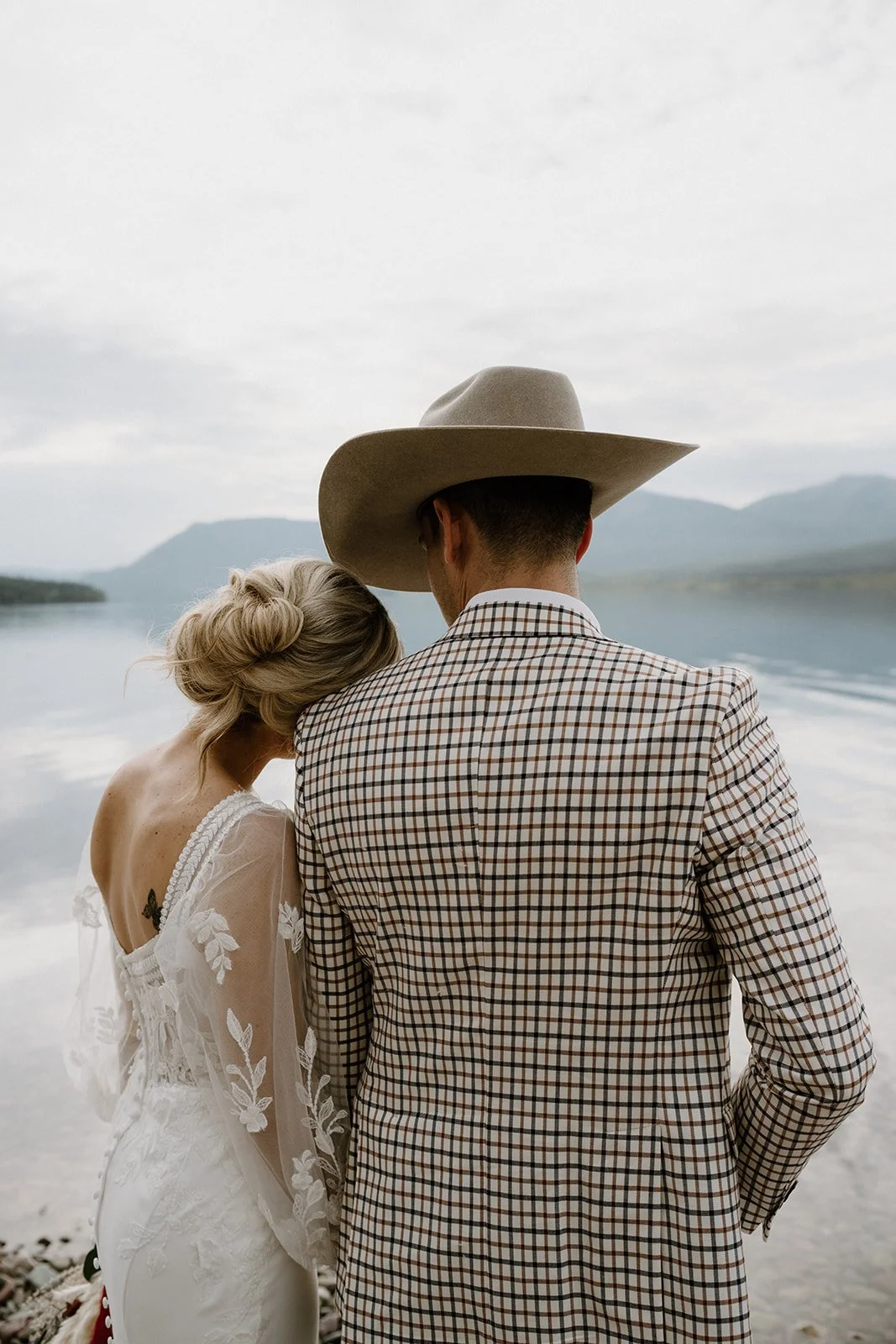 A couple, a woman in a white lace dress with her hair in a loose bun and a man in a plaid blazer and cowboy hat, stand close by a lake with mountains in the background on a cloudy day.