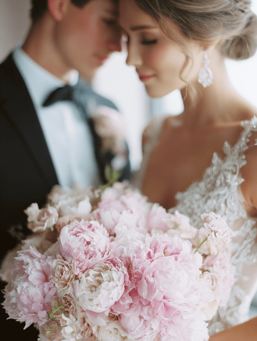 A bride and groom on their wedding day, close together with foreheads touching, holding a bouquet of pink flowers.