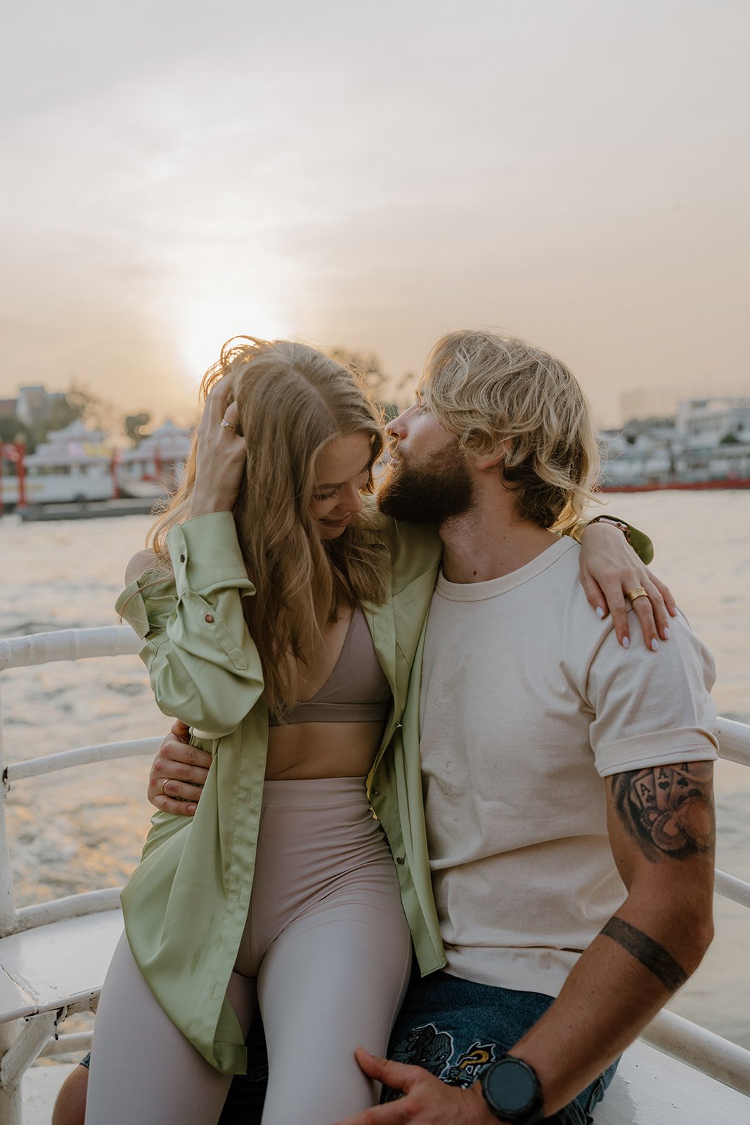 A couple sitting closely together near a waterfront at sunset, with boats and buildings in the background, sharing an intimate moment.