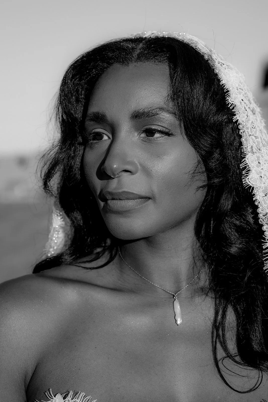 Black and white portrait of a woman with dark hair, wearing a lace veil, a necklace with a pendant, and a strapless top, outdoors.