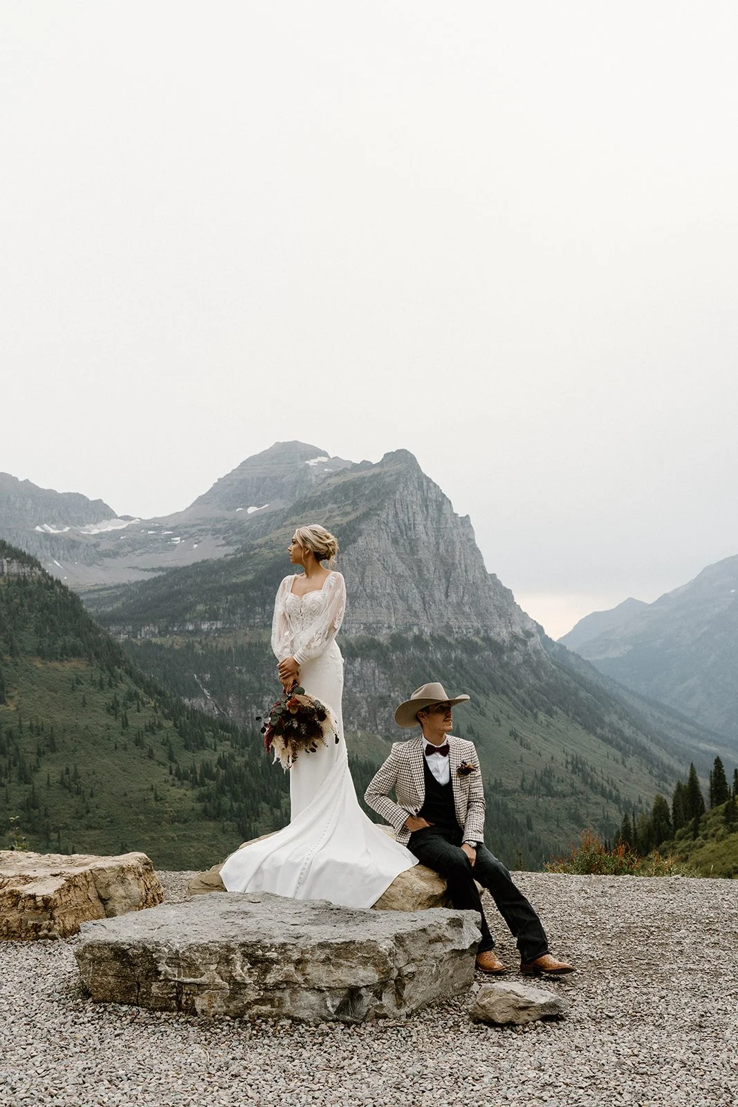 A bride in a white wedding dress holding a bouquet standing on a rock next to a man sitting on the same rock, in a mountainous landscape with mountains and trees in the background.