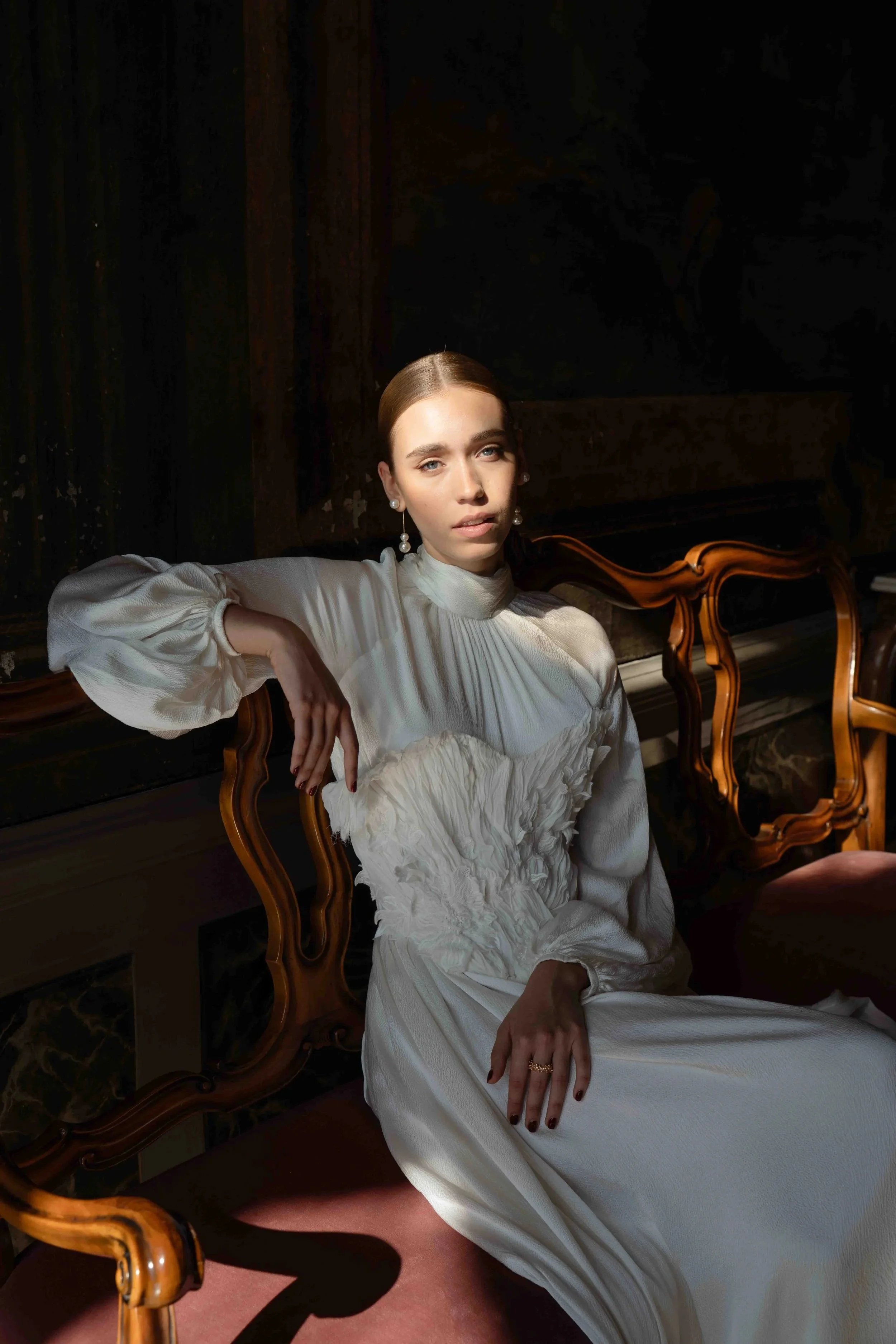 A young woman with pale skin and blue eyes sitting on a vintage wooden chair, wearing a long white dress with puffed sleeves and pearl earrings, in a dimly lit room with dark wooden walls and a piano in the background.