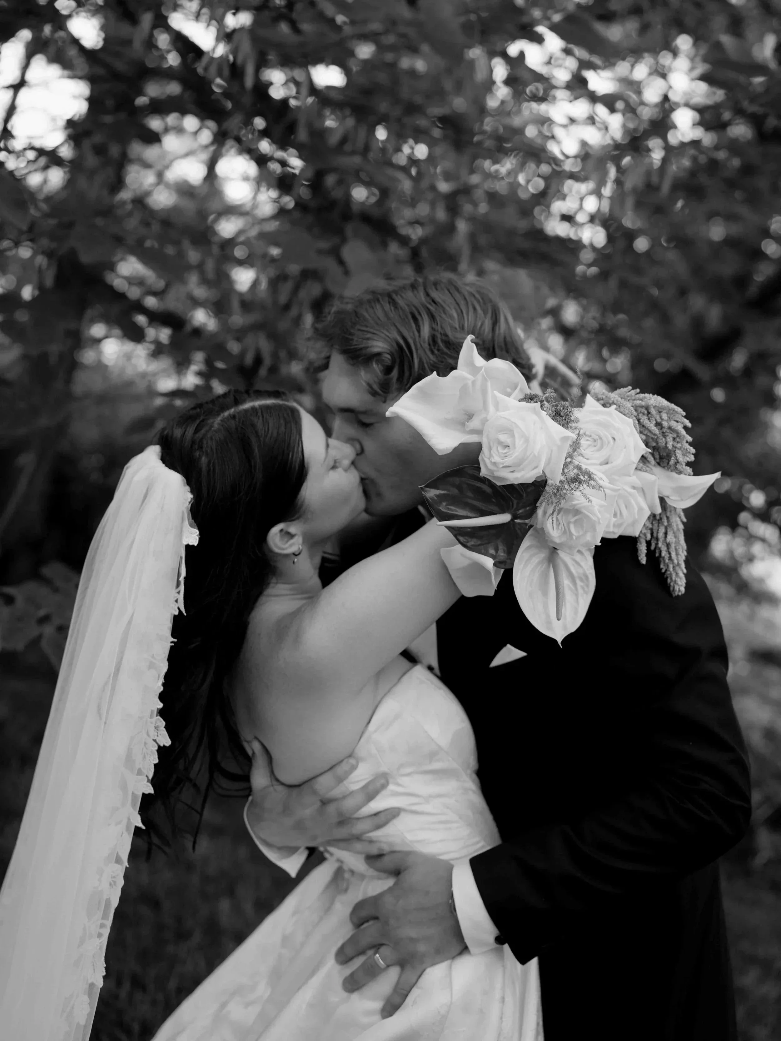 A black and white photo of a bride and groom sharing a kiss outdoors, the bride wearing a veil and holding a bouquet of large roses, the groom in a suit with a boutonniere.