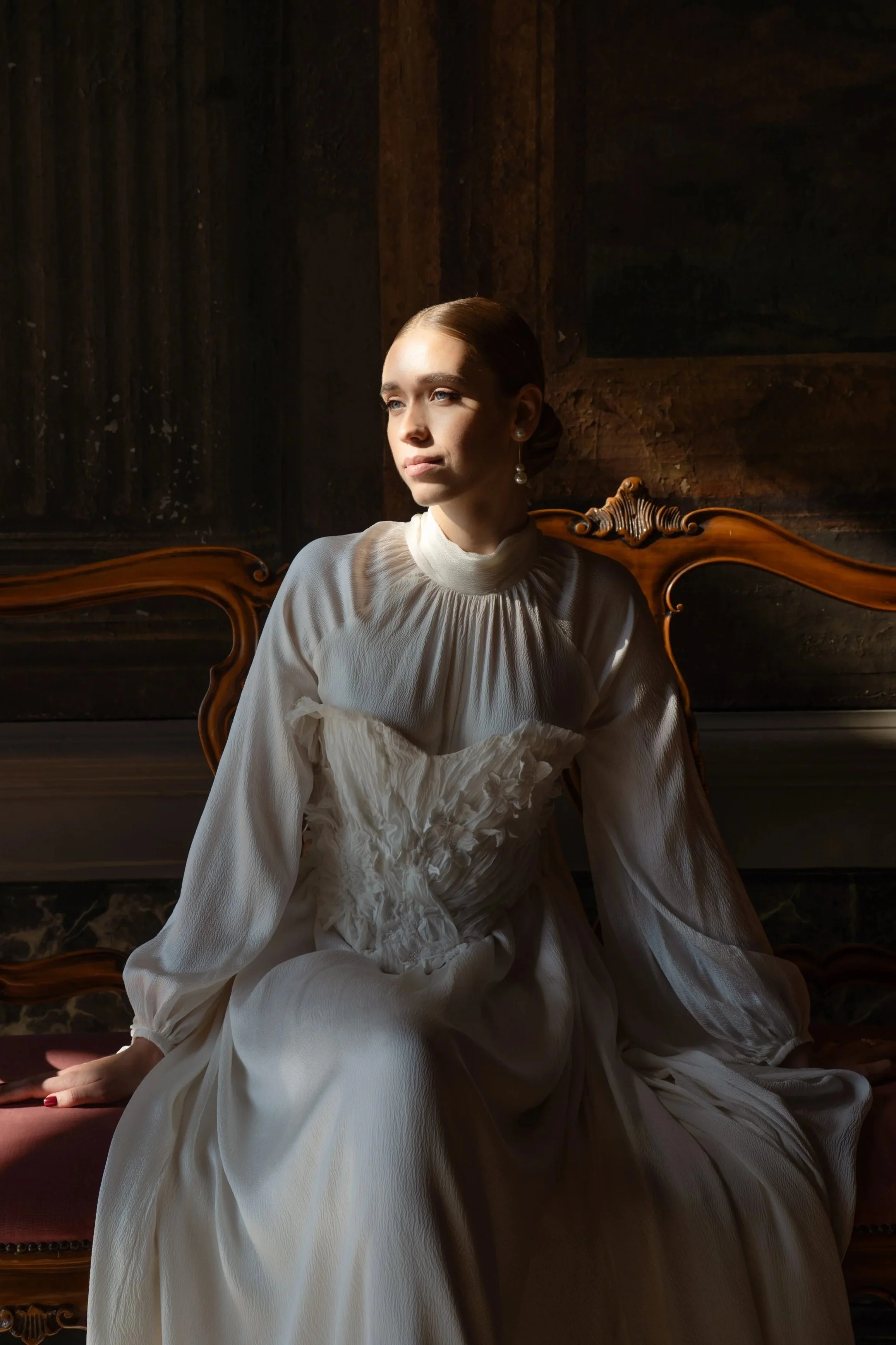 A woman with light skin and light-colored eyes, wearing a white vintage-inspired dress with puffed sleeves and a high ruffled collar, sitting on a vintage wooden bench with an ornate backrest, and looking to the left. She has her hair styled in a sleek bun, accessorized with pearl earrings, in a softly lit setting with a dark wooden background.