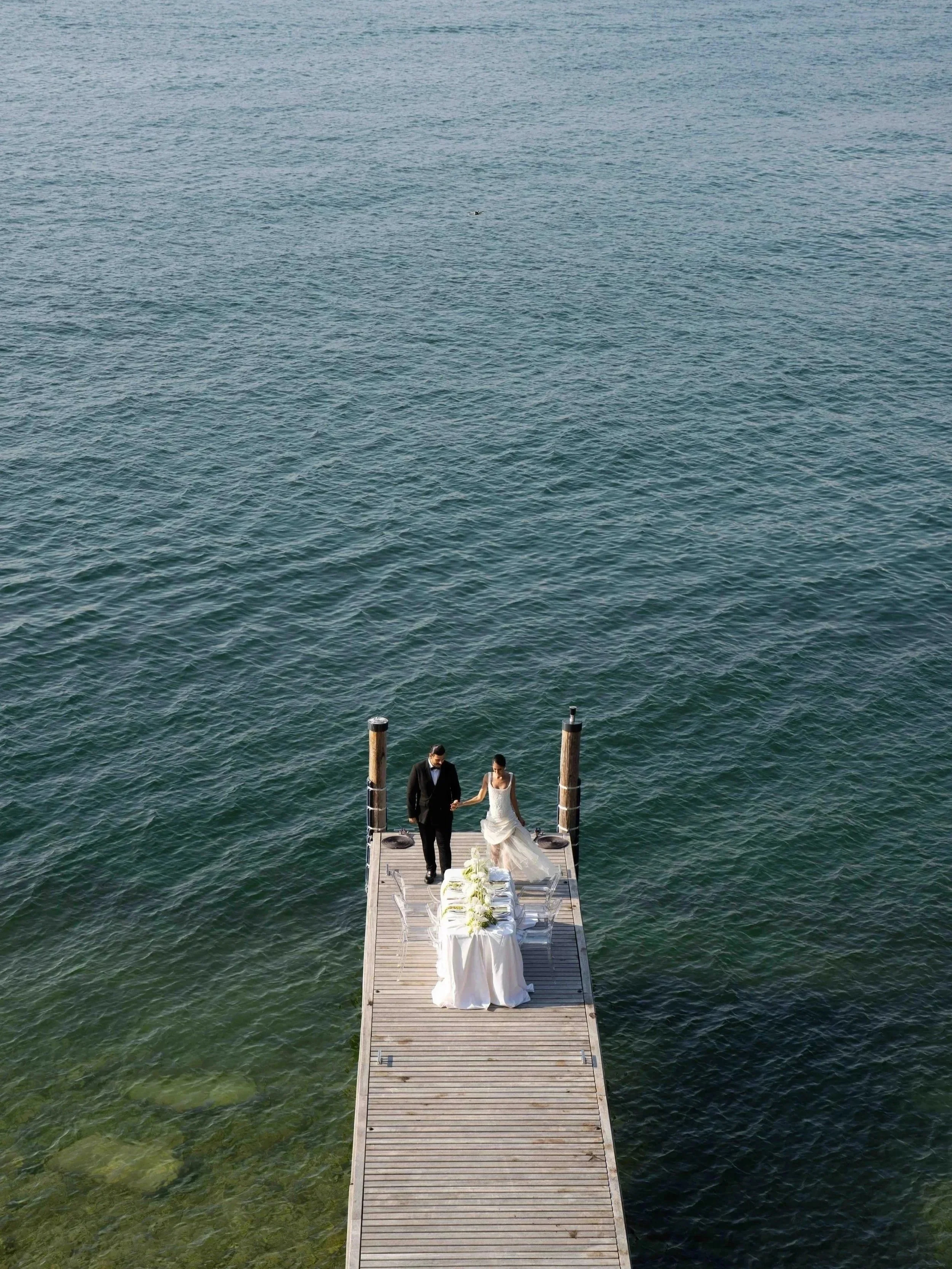 A bride and groom walking hand in hand on a wooden dock over the water, dressed in wedding attire, with tables set up on the dock for a wedding celebration.
