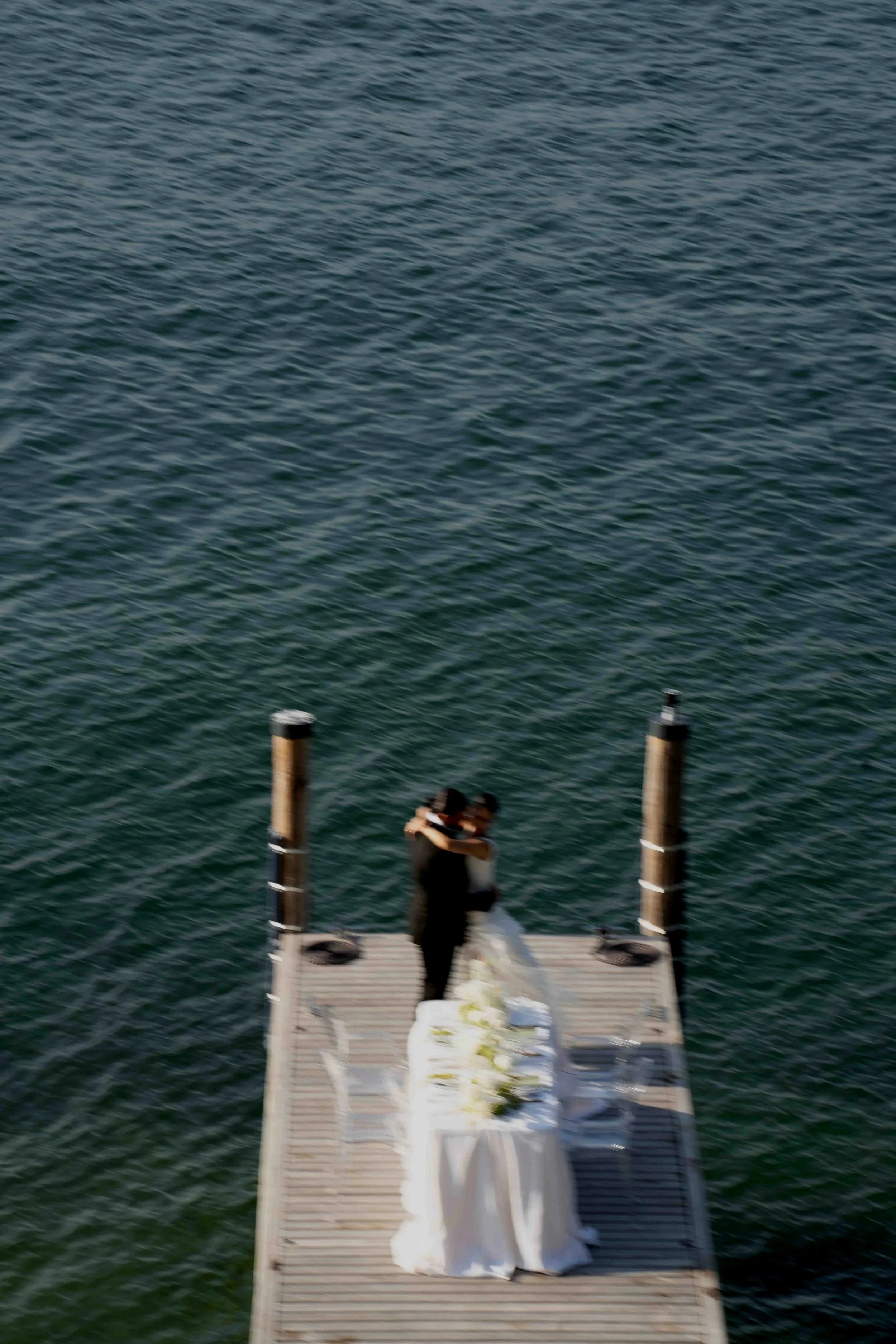 A couple in wedding attire embracing on a dock by the water, with wedding flowers and decorations nearby.