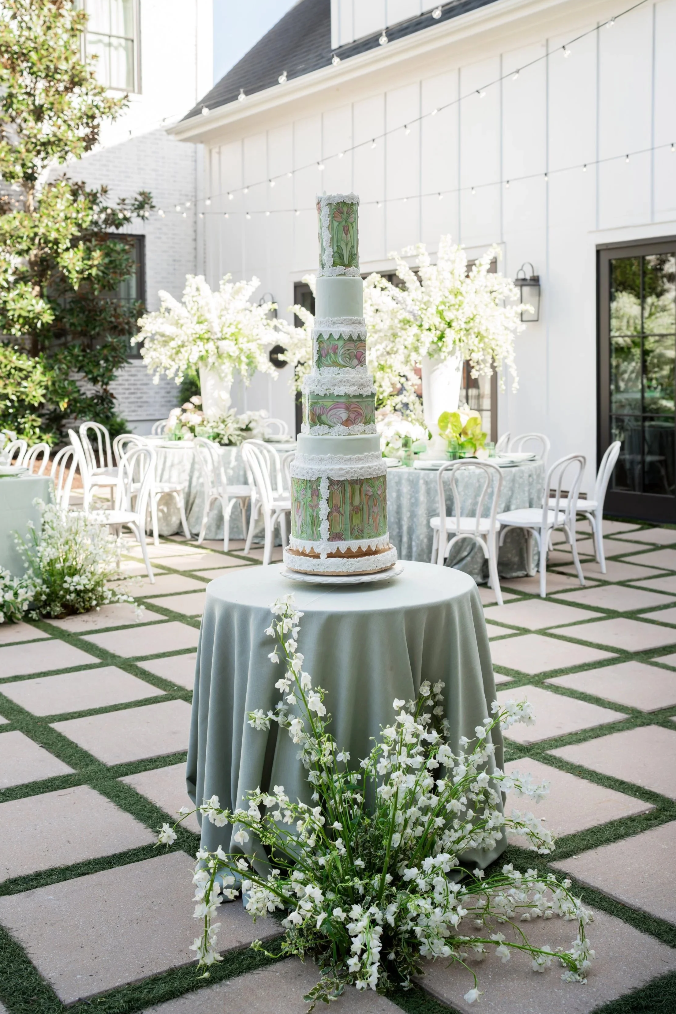 A tall cake decorated with floral patterns and lace, placed on a round table with a gray tablecloth, surrounded by white flowers and greenery, set in an outdoor garden with white chairs, floral arrangements, and string lights, under a partly cloudy sky.