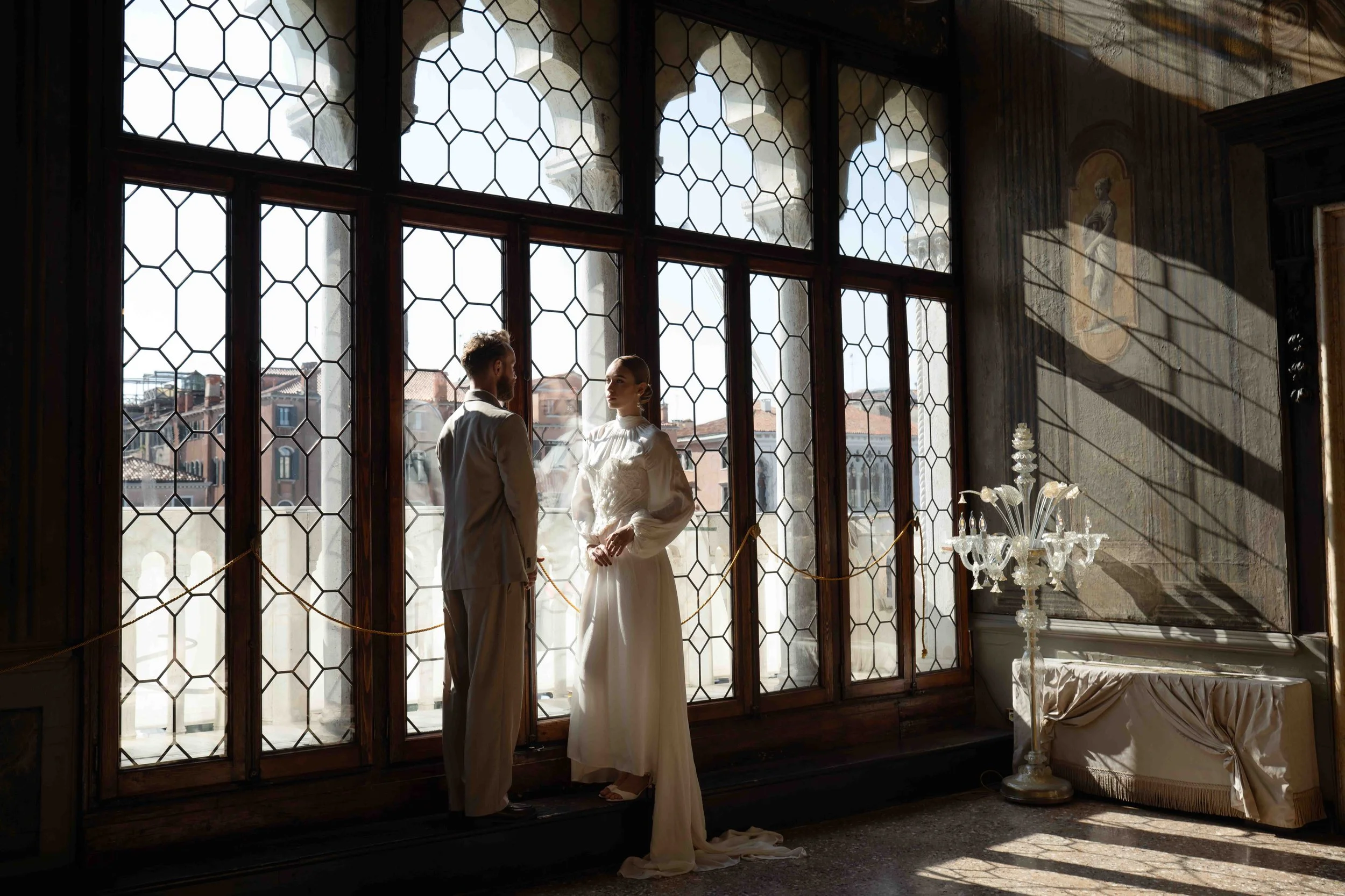 A bride and groom standing inside a historic building with large, ornate windows and decorative details, possibly during a wedding or engagement.