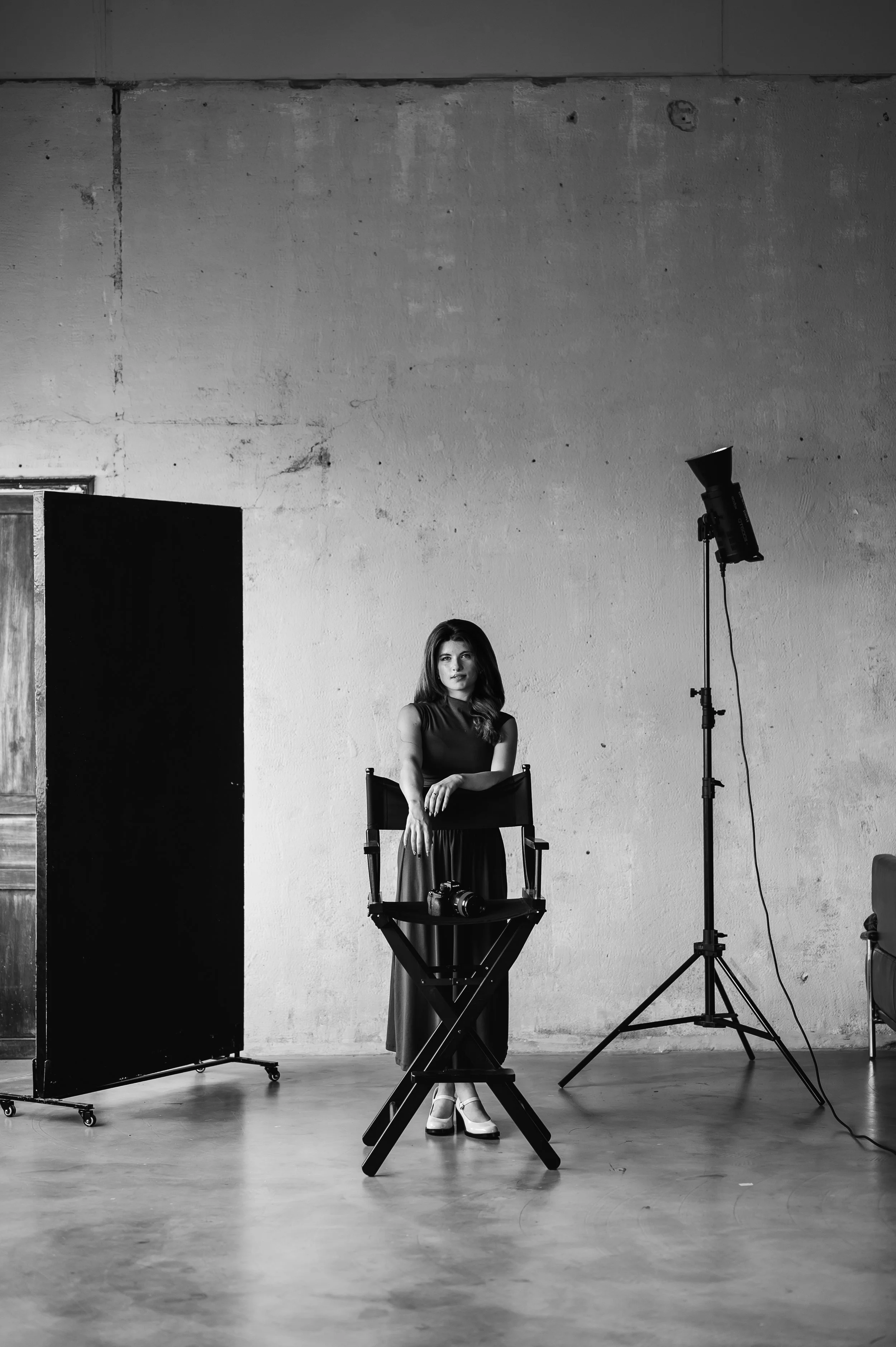 A woman in a long skirt and high heel shoes sitting on a director's chair in a photography studio with a large softbox light to her right, a backdrop to her left, and a camera on a tripod in front of her. The studio has a distressed concrete wall.