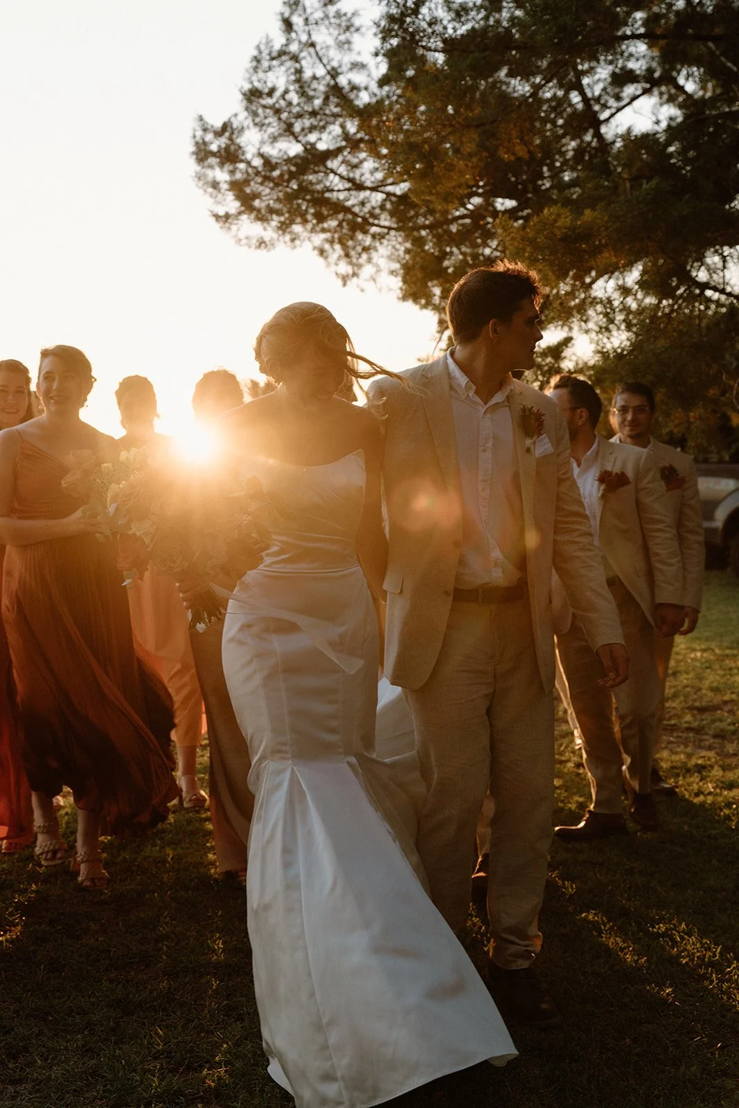 A group of wedding guests walking outdoors during sunset, with the bride and groom in the foreground. The bride is in a white wedding gown, and the groom in a light-colored suit, holding hands. The sunlight creates a warm glow and lens flare, with trees in the background.