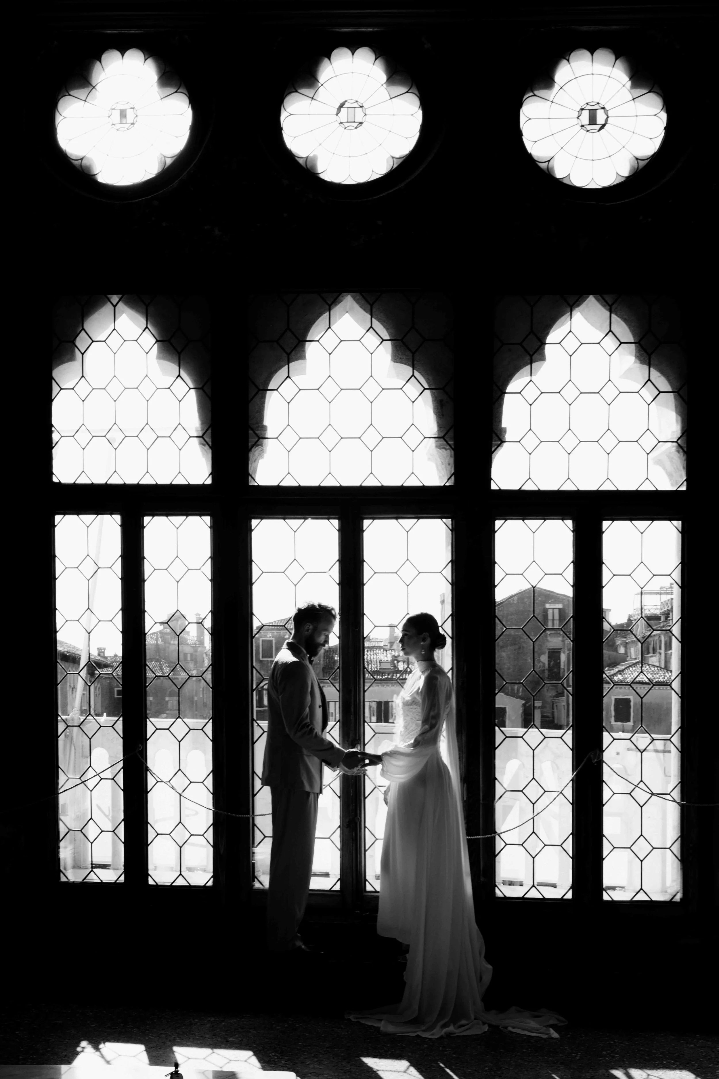 A black and white photo of a bride and groom holding hands and facing each other in front of large stained glass windows, with sunlight streaming through.